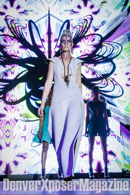 Runway model in a turquoise sculptural gown and futuristic jewelry walks before a vivid neon screen at the Alien Life fashion show.