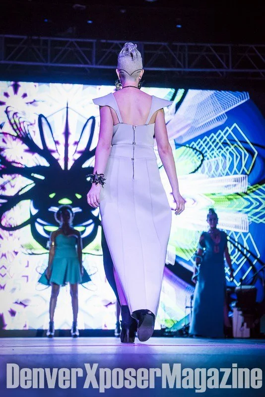 Runway model in a turquoise sculptural gown and futuristic jewelry walks before a vivid neon screen at the Alien Life fashion show.