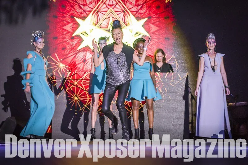 Runway model in a turquoise sculptural gown and futuristic jewelry walks before a vivid neon screen at the Alien Life fashion show.