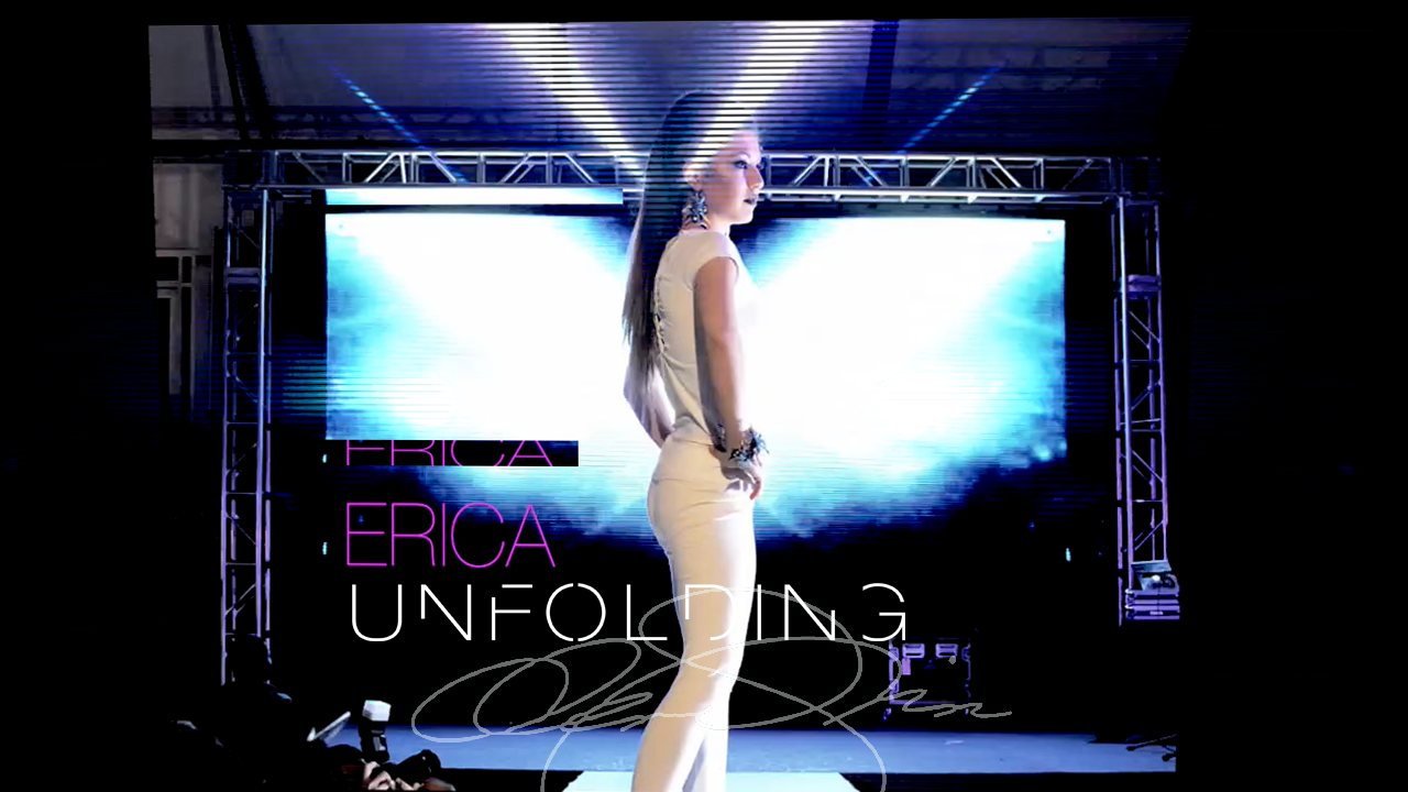 Runway model in a white fitted outfit wears statement earrings and a crystal cuff before a blue-lit screen titled “Erica Unfolding.”