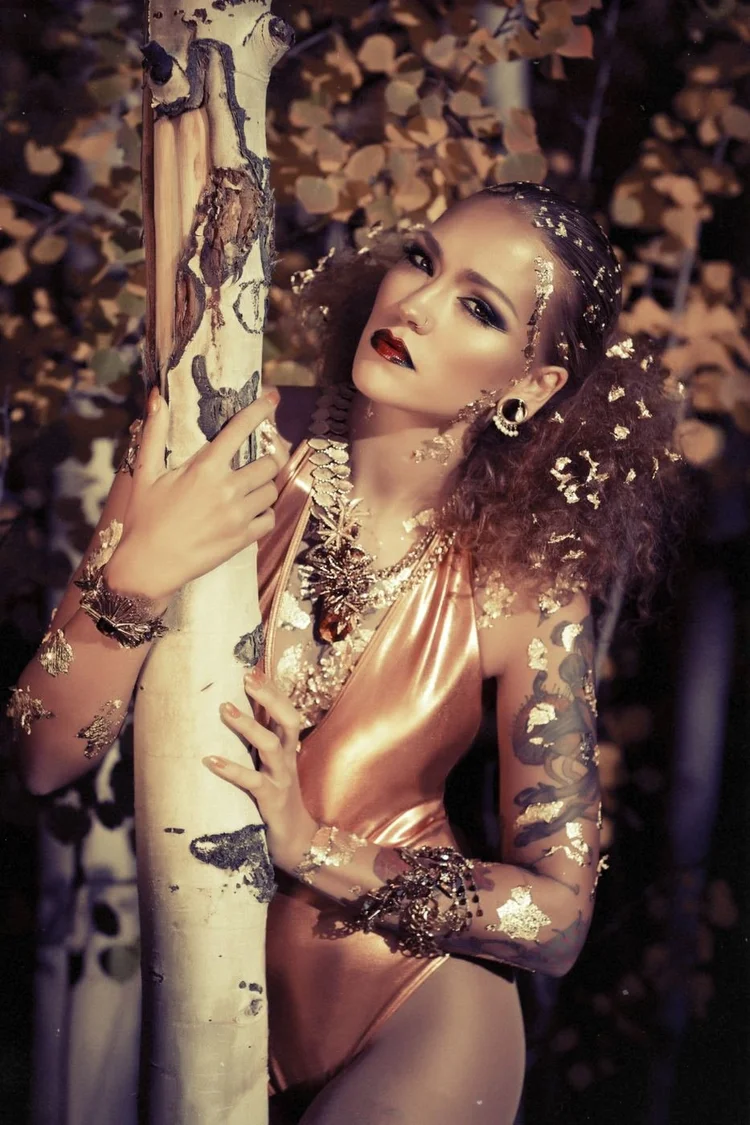 Model Stefanie Dolenc with gold leaf on her skin, curly hair and Andrea Li gold and crystal pieces, against an aspen trunk