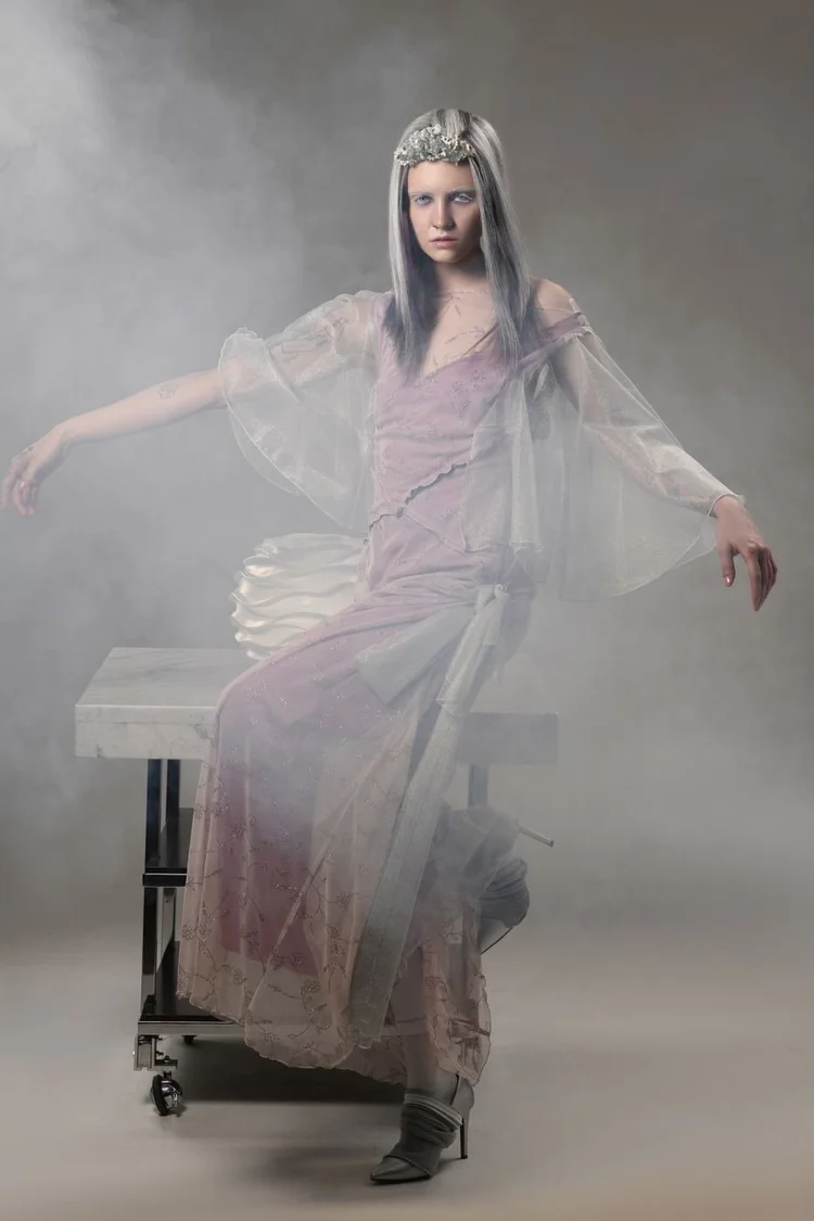 A model in a sheer lilac gown and headpiece seated on an industrial table