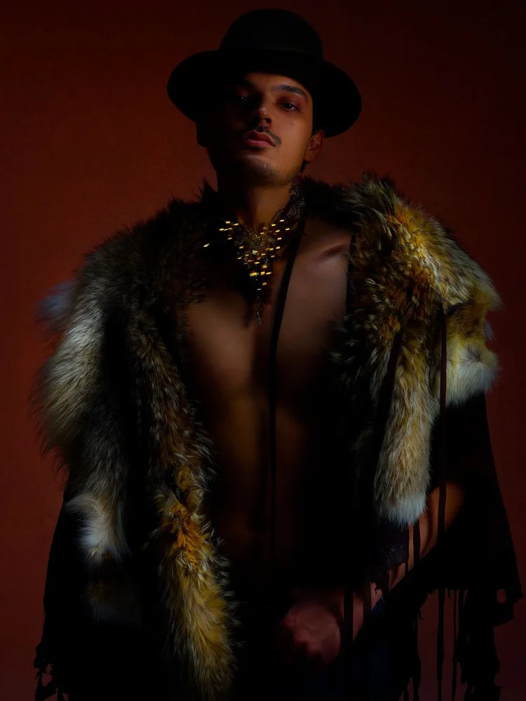 A man in a black bowler hat and a fox fur wrap over a bare chest, wearing an Andrea Li gold spiked cluster collar, on a rust ground