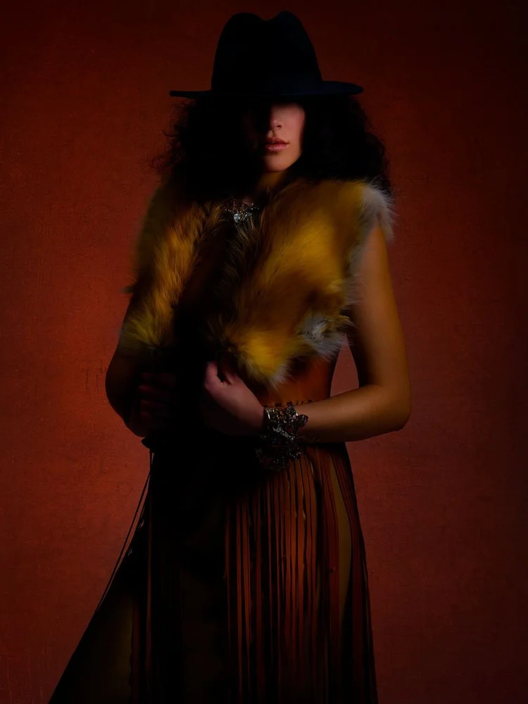 A woman in a black fedora and golden fur stole over a fringed leather skirt, with an Andrea Li crystal cluster cuff