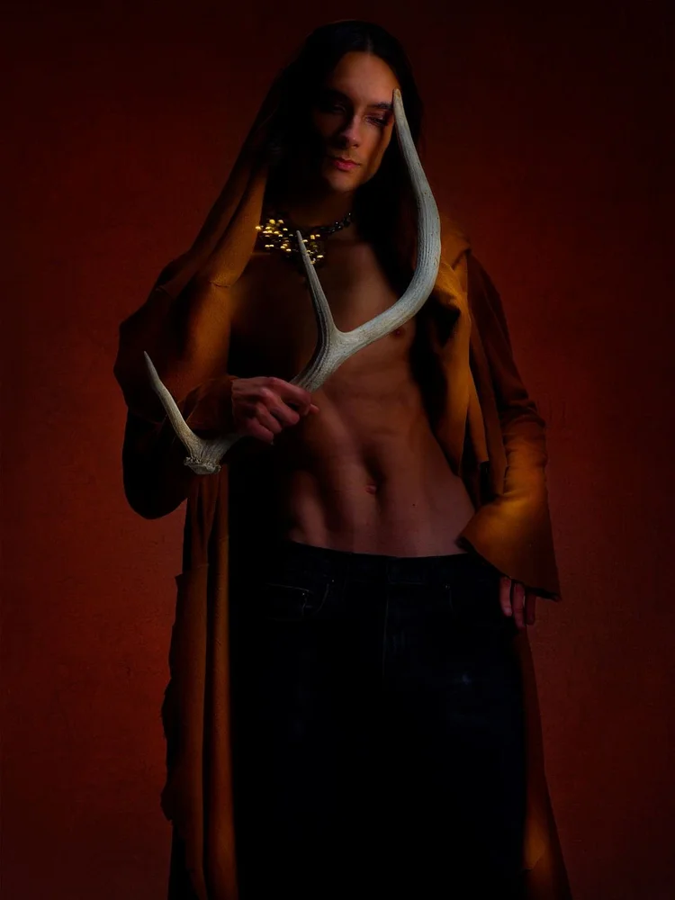 A long-haired man in a hooded suede robe holding an antler, with an Andrea Li gold cluster collar
