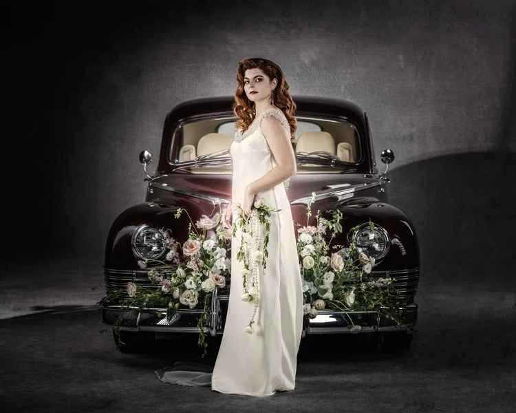 A bride in an ivory gown with a cascading bouquet before a vintage car
