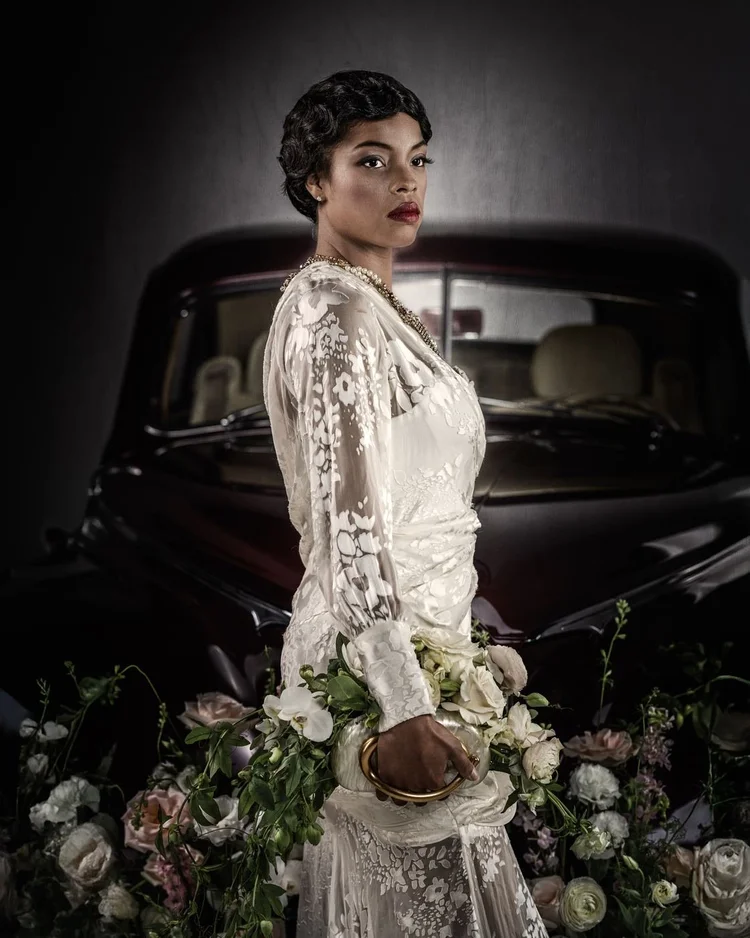 A model in a lace gown in profile beside the car, bouquet in hand