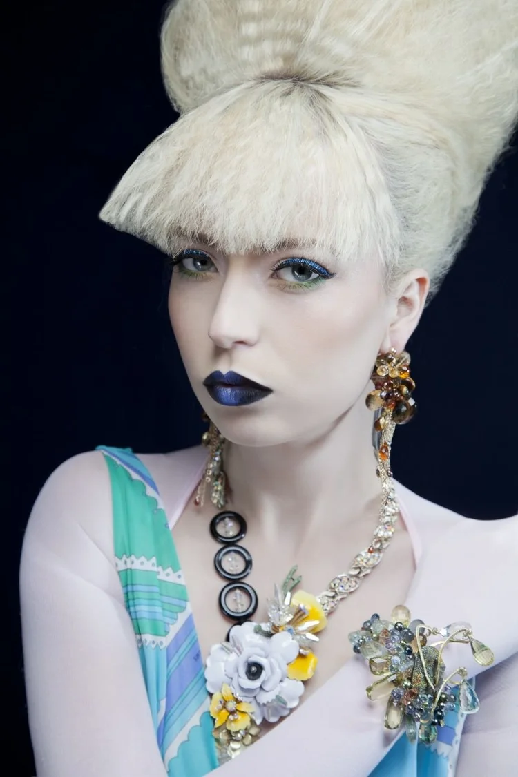 Close portrait: a crimped platinum beehive, blue lips and an Andrea Li floral and ring cluster necklace over a pastel print top
