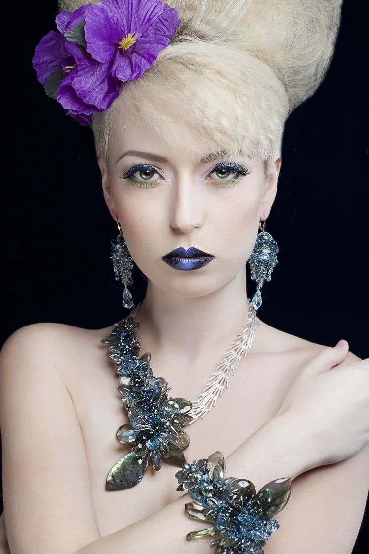A platinum beehive with a purple flower, blue lips, arms crossed, wearing Andrea Li blue crystal cluster earrings, necklace and cuff