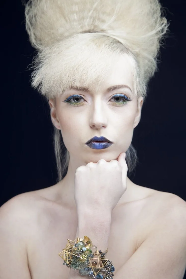 A platinum beehive and blue lips, chin resting on her fist, with an Andrea Li gold geometric and gemstone cluster cuff