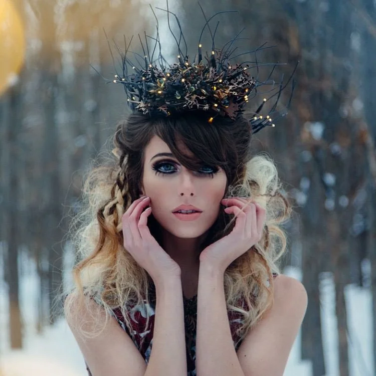 A model in a snowy forest wearing a crown of black twigs and tiny lights, hands at her face