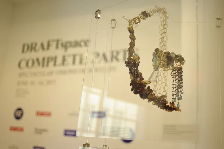 An Andrea Li cluster necklace on a clear panel in front of the DRAFTspace entrance wall