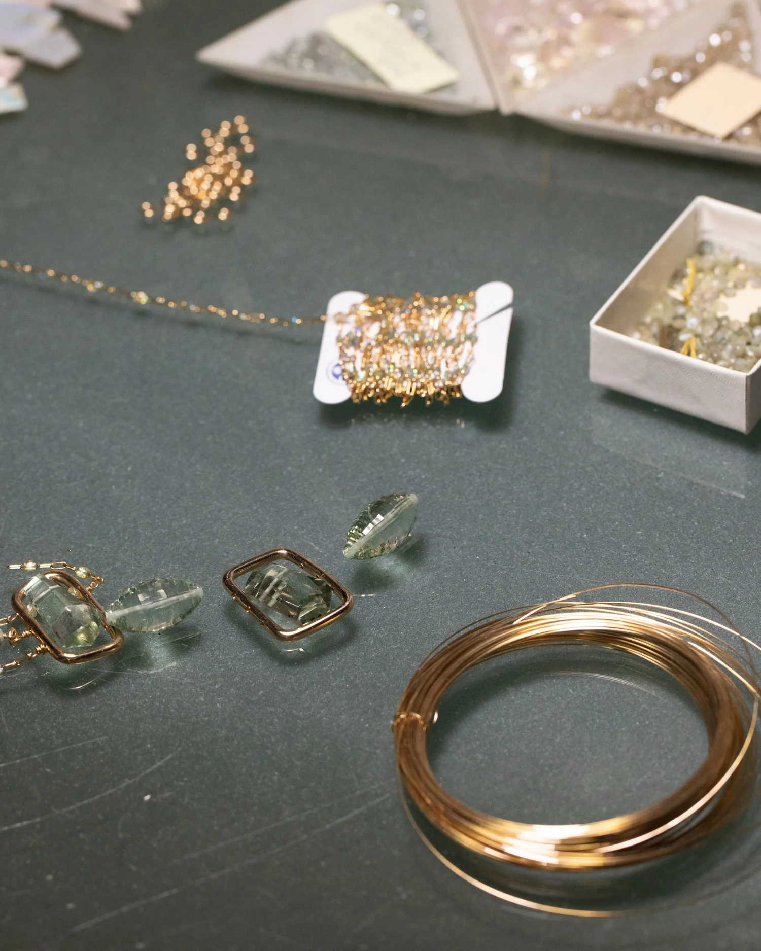 On Andrea Li's bench: green gemstones set in gold frames, a coil of gold wire and a spool of gemstone chain