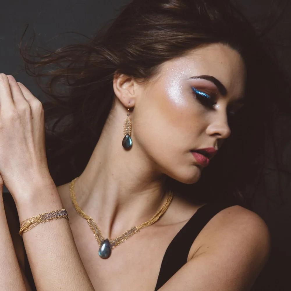 A model in a black top wearing Andrea Li gold drop earrings with a dark teardrop gemstone, a matching pendant and a gold bracelet