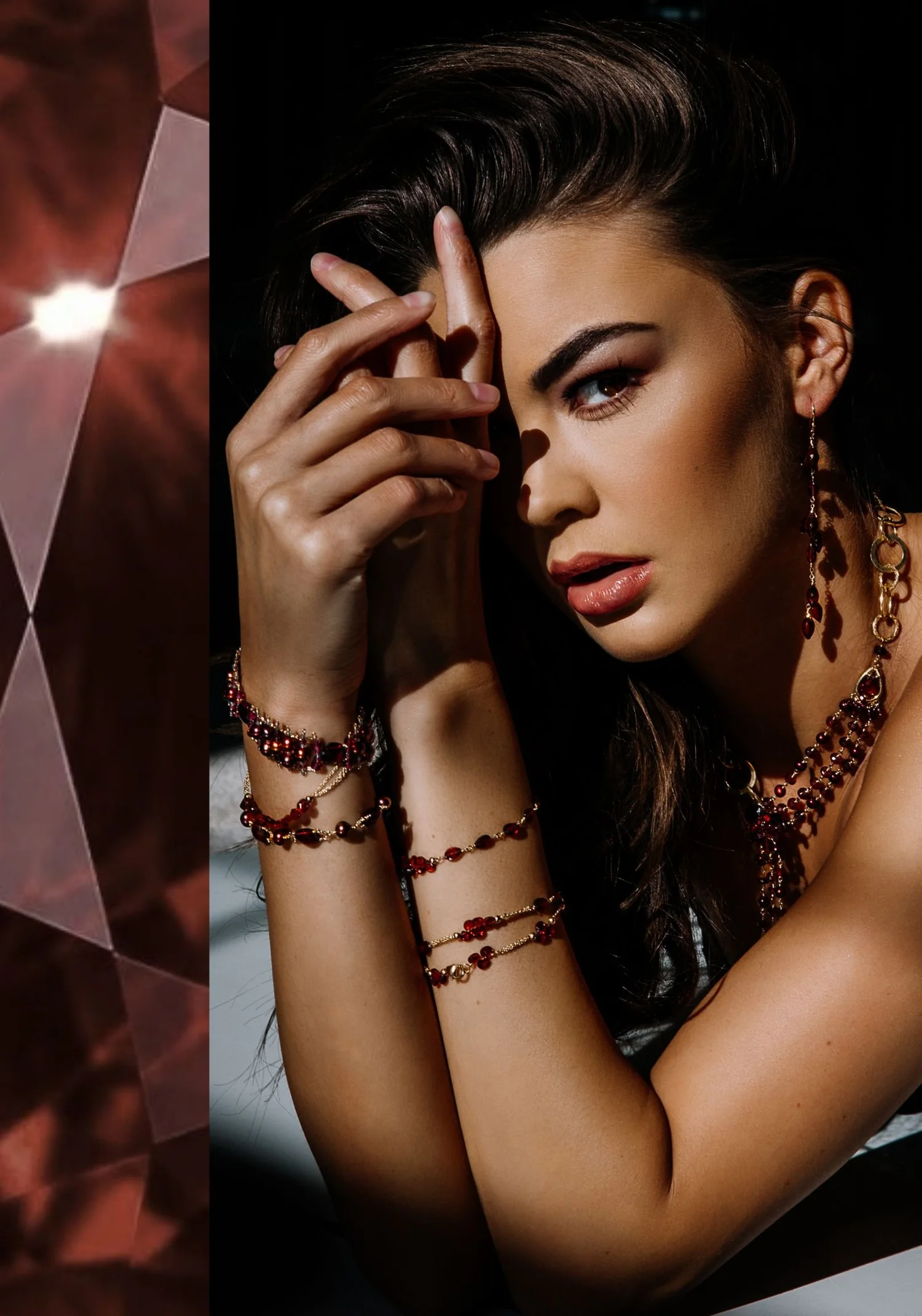 A model with dark swept-back hair, a hand at her brow, wearing a garnet and gold multi-strand necklace, long garnet drop earrings and stacked garnet bracelets, beside a close view of a faceted garnet