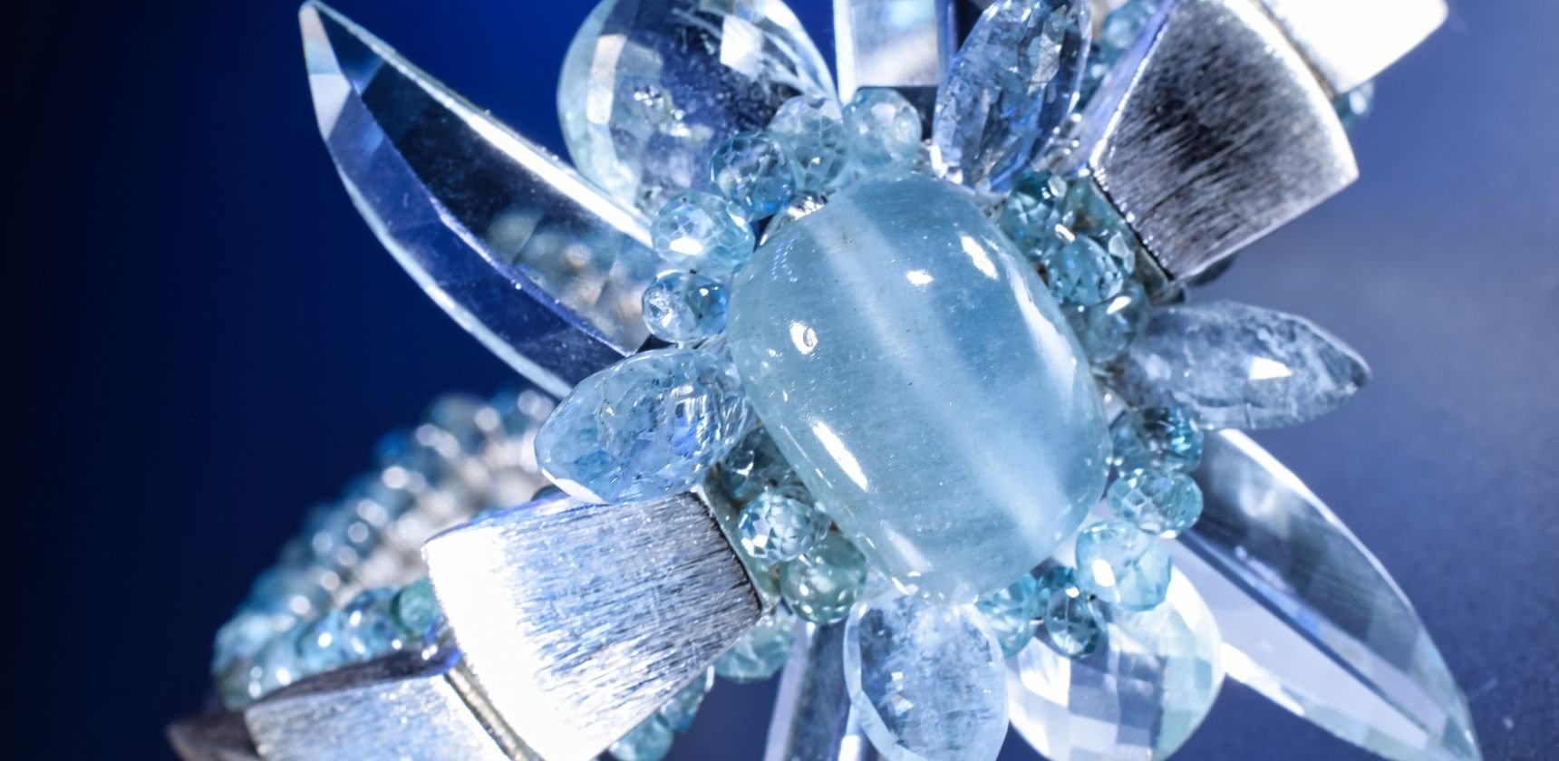 Close view of an Andrea Li statement cuff: a milky aquamarine cabochon framed by brushed silver petals and faceted blue gemstones on deep blue