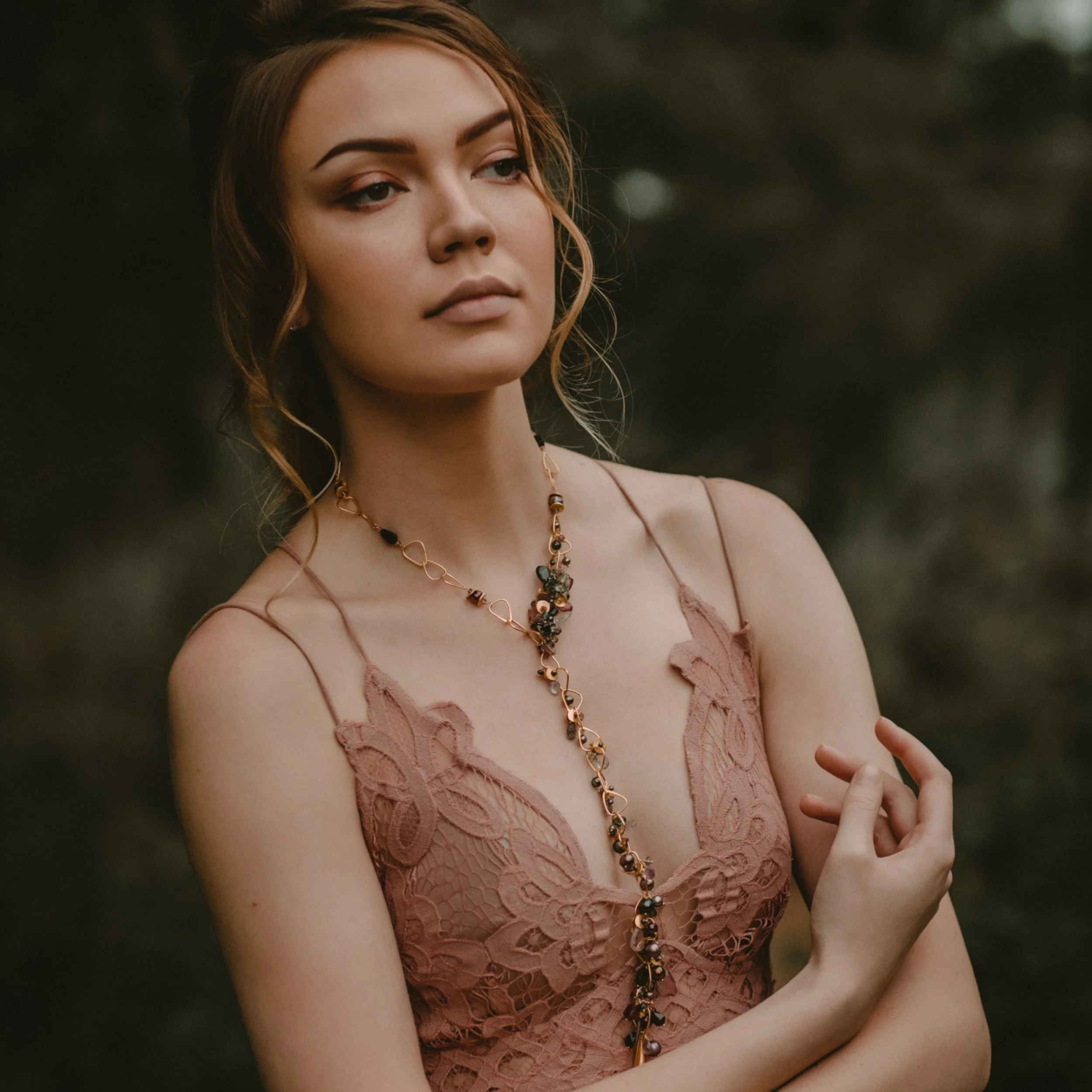 Portrait of a woman in a blush lace dress wearing a delicate drop necklace, posed against a softly blurred woodland background.