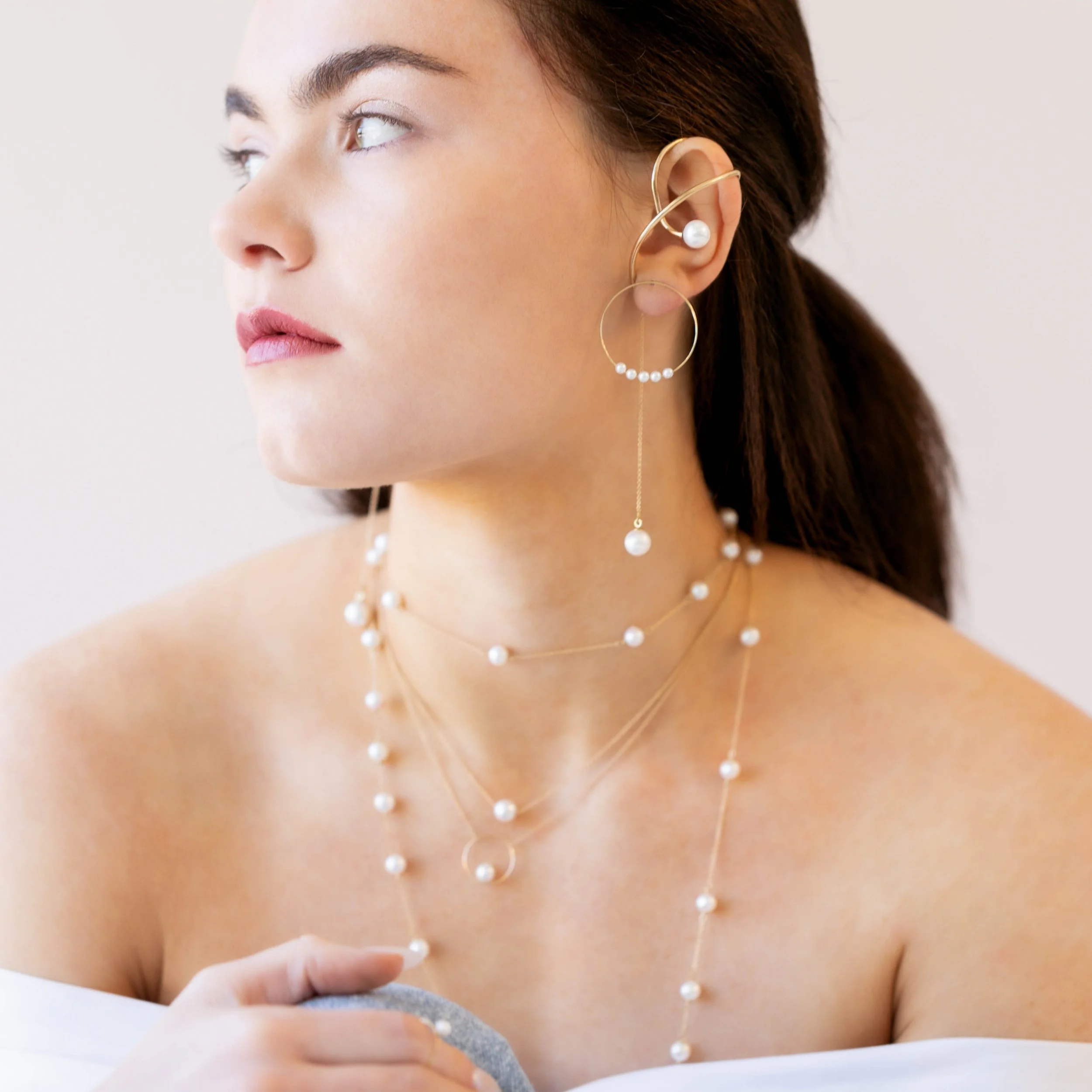 Portrait of a woman wearing layered pearl jewelry and a sculptural gold ear cuff, posed against a soft neutral background.