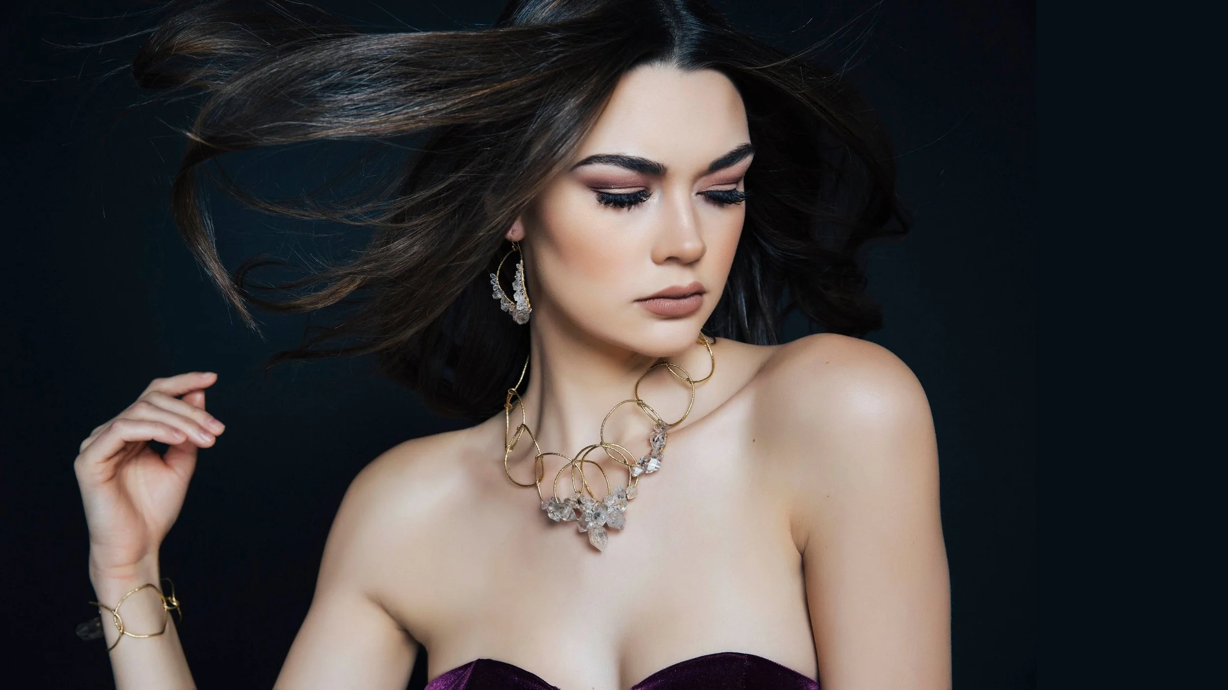 Editorial portrait of a model wearing sculptural gold and crystal jewelry, with flowing dark hair against a dramatic black background.
