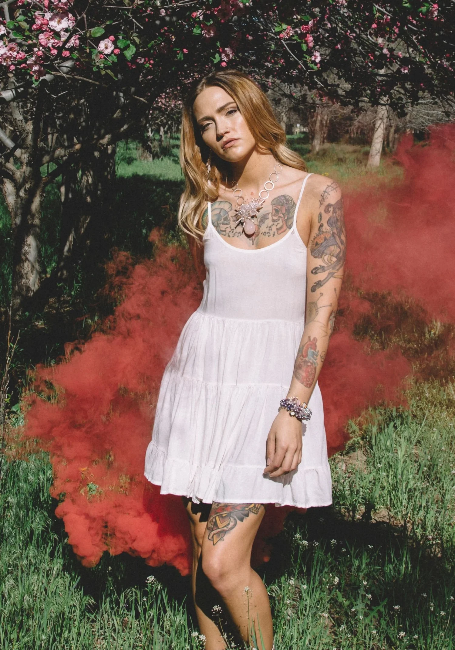 A tattooed model in a white sundress in a blossoming orchard, red smoke billowing beside her, wearing a pale crystal statement necklace and a gemstone bracelet