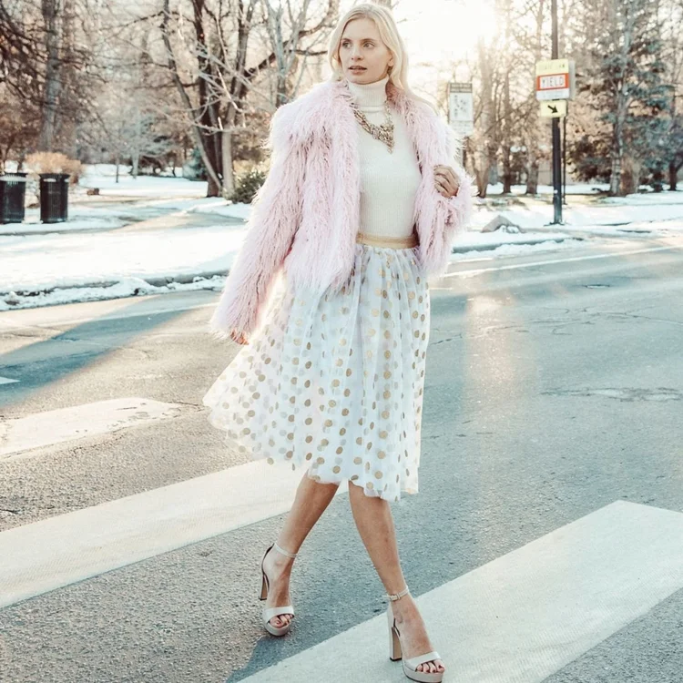Model in a pink fur coat and a white polka dot skirt crossing a snowy street, wearing a statement necklace