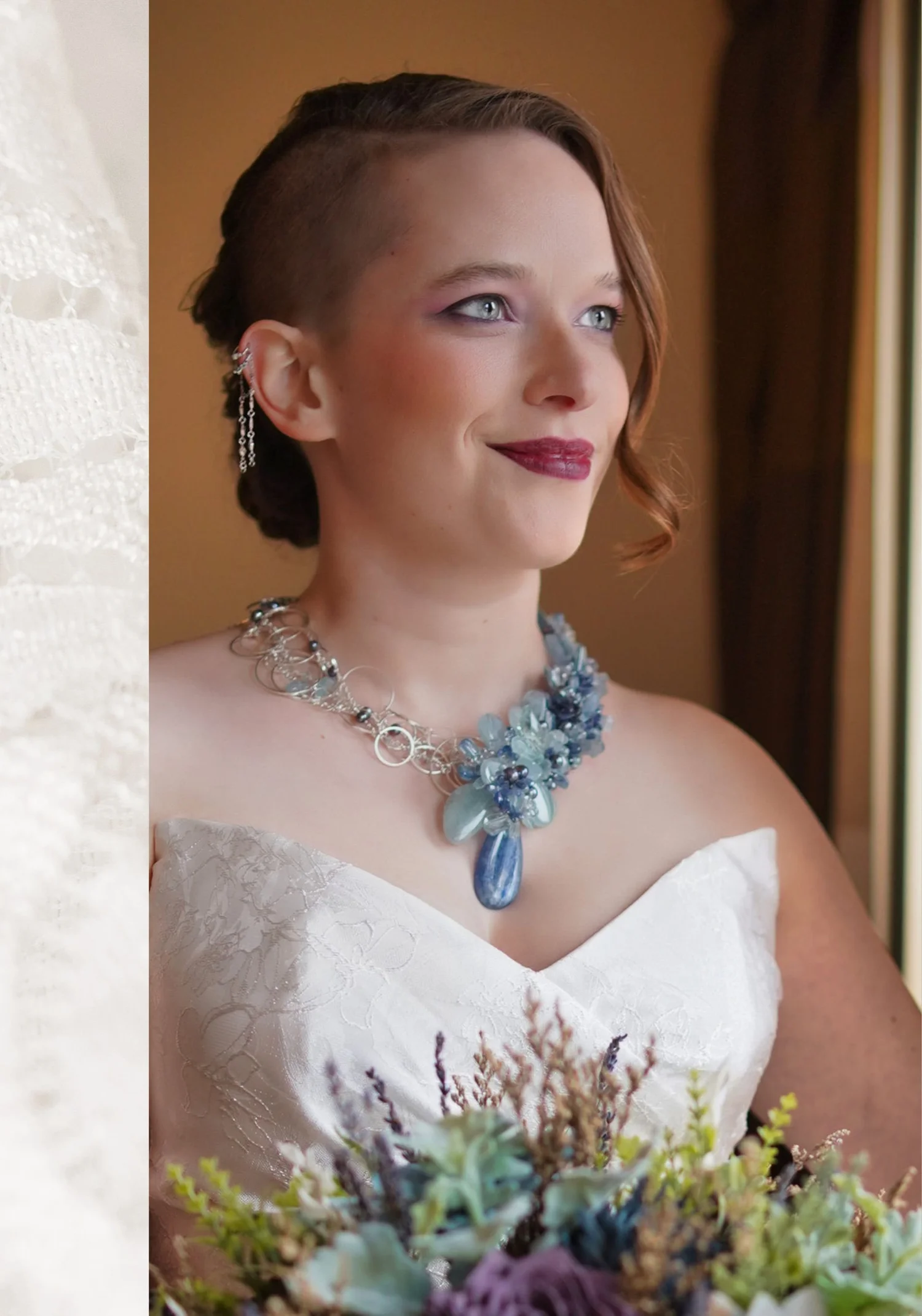 A bride in a strapless lace gown wearing a silver wire necklace with a cluster of blue gemstones and a blue drop, and a silver chain ear cuff, holding a bouquet of succulents and lavender