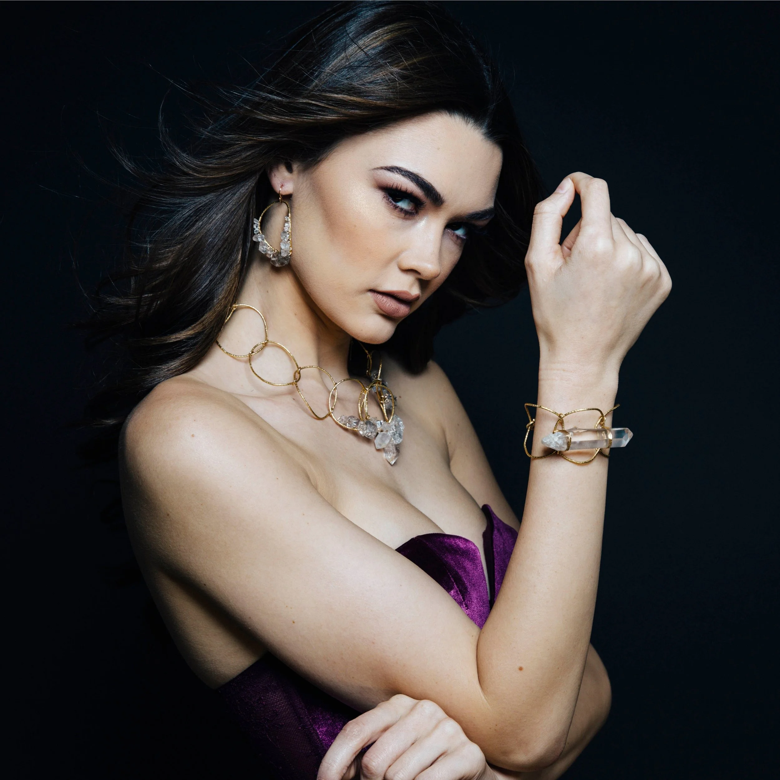 Portrait of a model in a purple velvet dress wearing sculptural gold jewelry with clear crystals against a dark background.