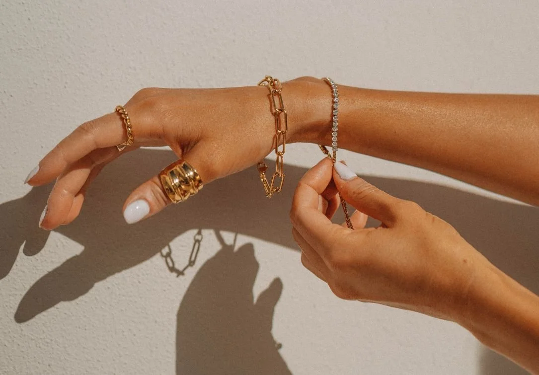Close-up of a hand wearing layered gold bracelets and rings while fastening a delicate tennis bracelet against a neutral wall.