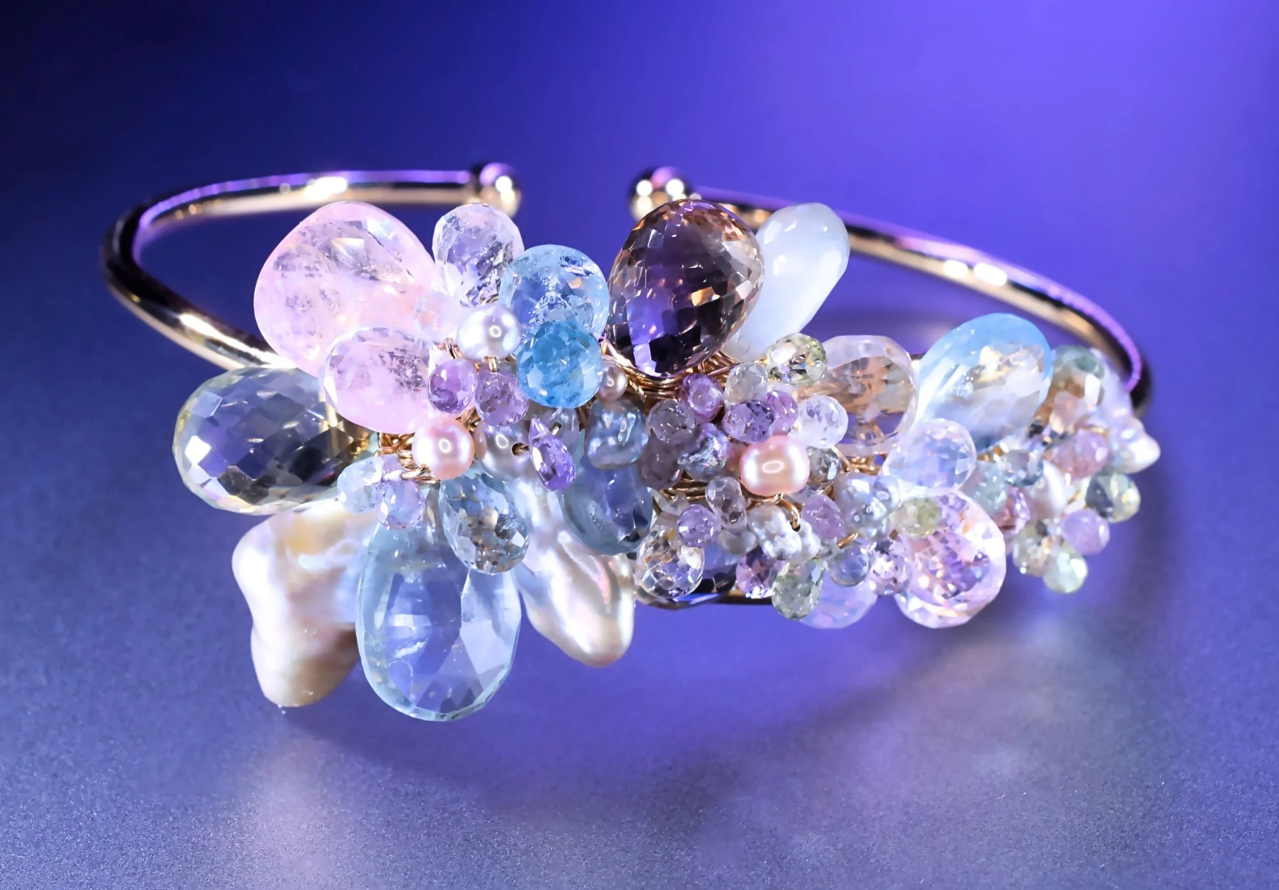 Close-up of a gold cuff bracelet adorned with pastel gemstones and pearls, set against a luminous deep blue background.