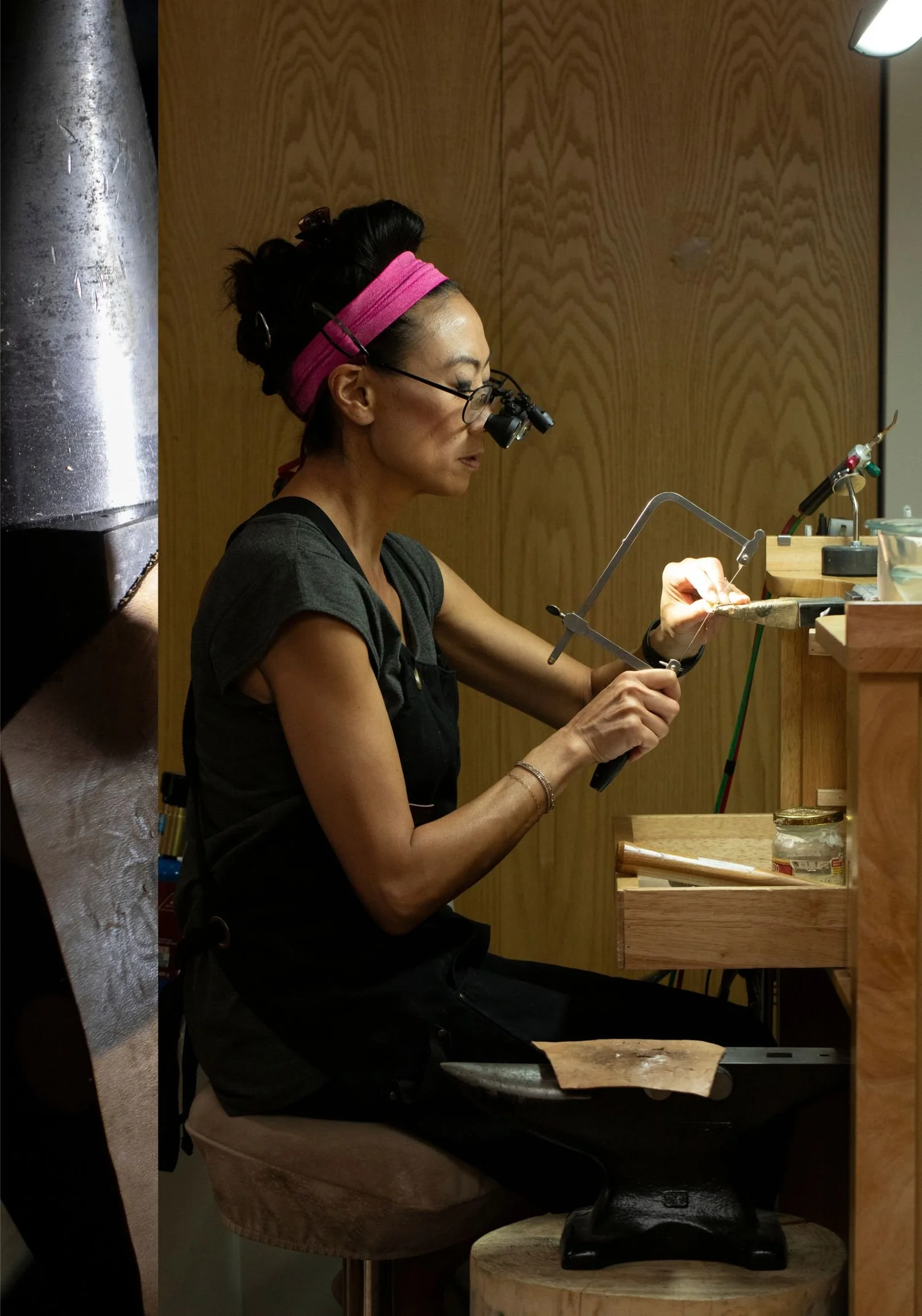 Andrea Li at her jeweler's bench in a pink headband and magnifying loupes, sawing metal with a jeweler's saw