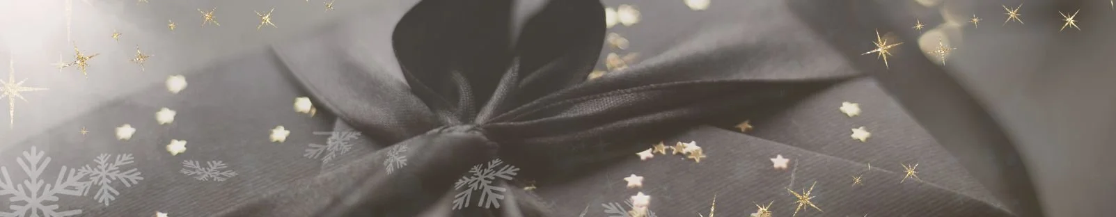 Close-up of a wrapped gift with black ribbon, soft holiday bokeh, gold stars, and snowflake overlays in muted monochrome tones.