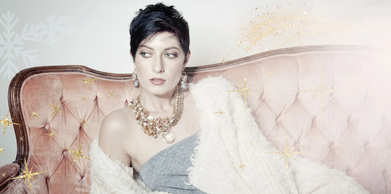 A model with short dark hair on a blush velvet settee, wearing large drop earrings and a gold statement necklace of champagne stones