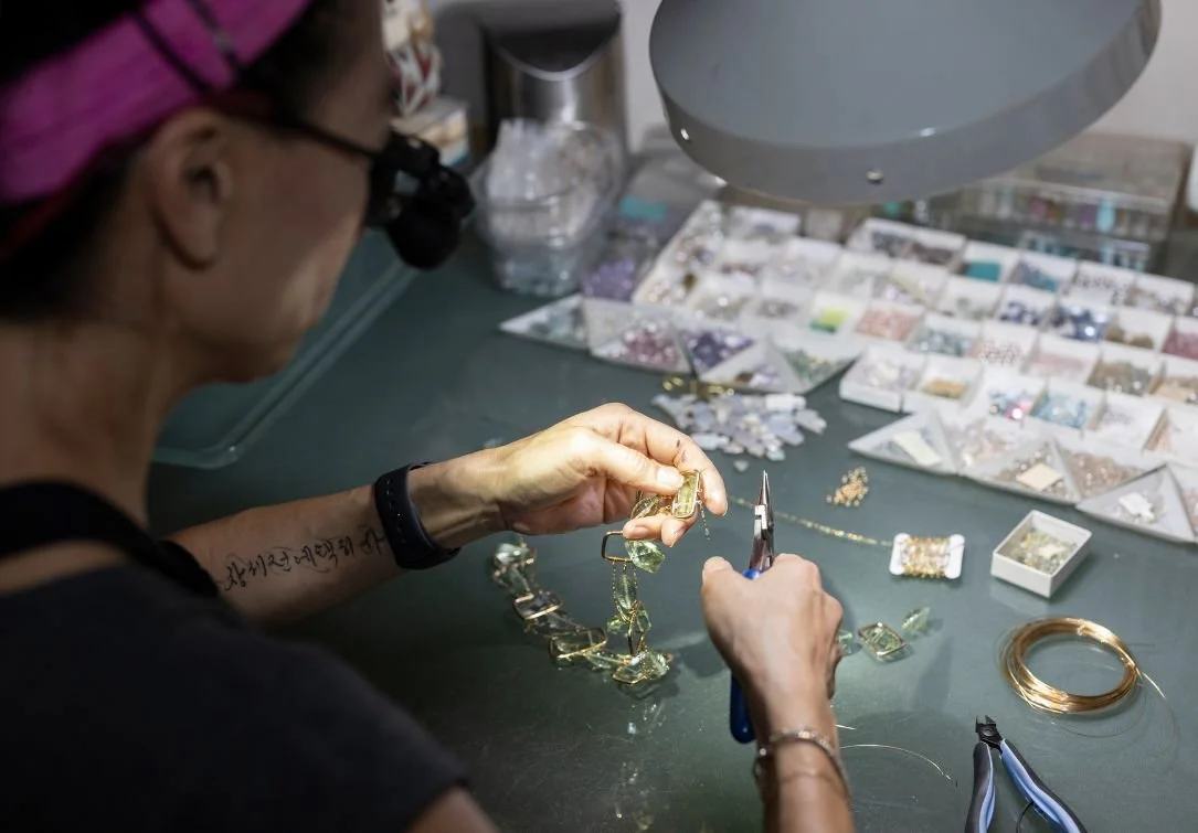 Jeweler at a workbench using pliers on a gemstone piece, surrounded by trays of beads, tools, and wire under a task lamp.