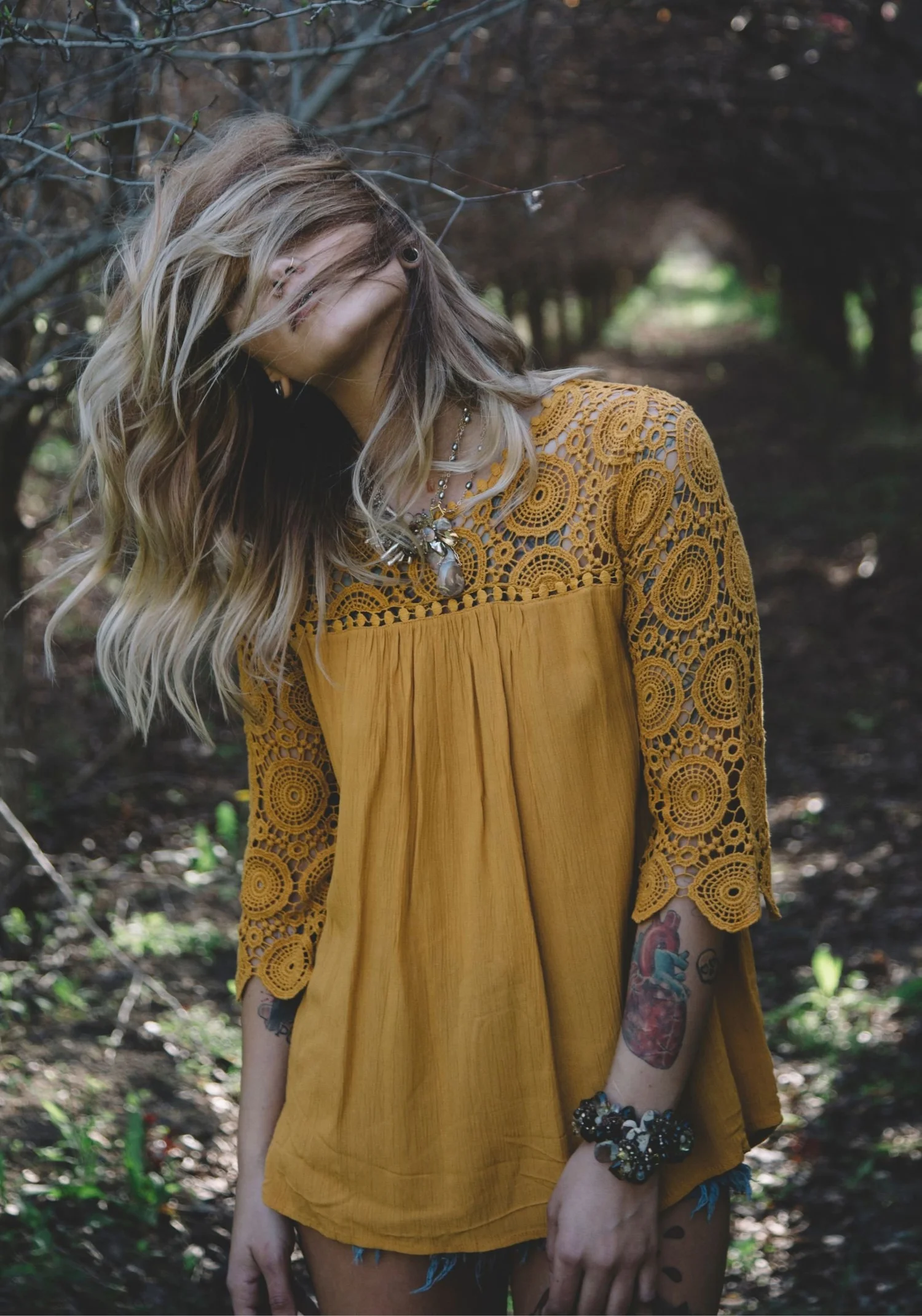 A model in a saffron lace top in a shaded orchard, hair across her face, wearing a gemstone pendant and a wide gemstone cuff