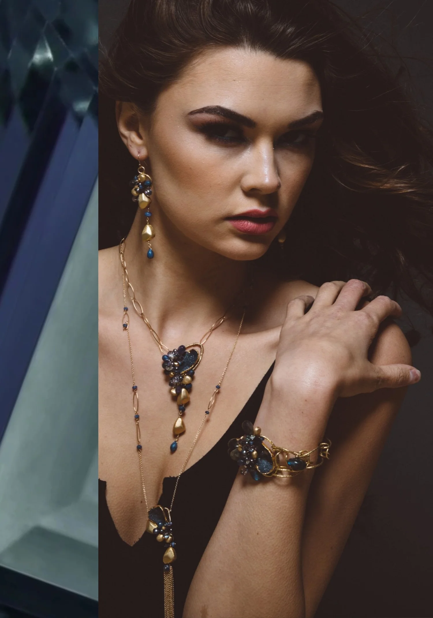 A model wearing long drop earrings, a layered gold chain necklace and a gold link bracelet, all clustered with deep blue gemstones and gold nuggets