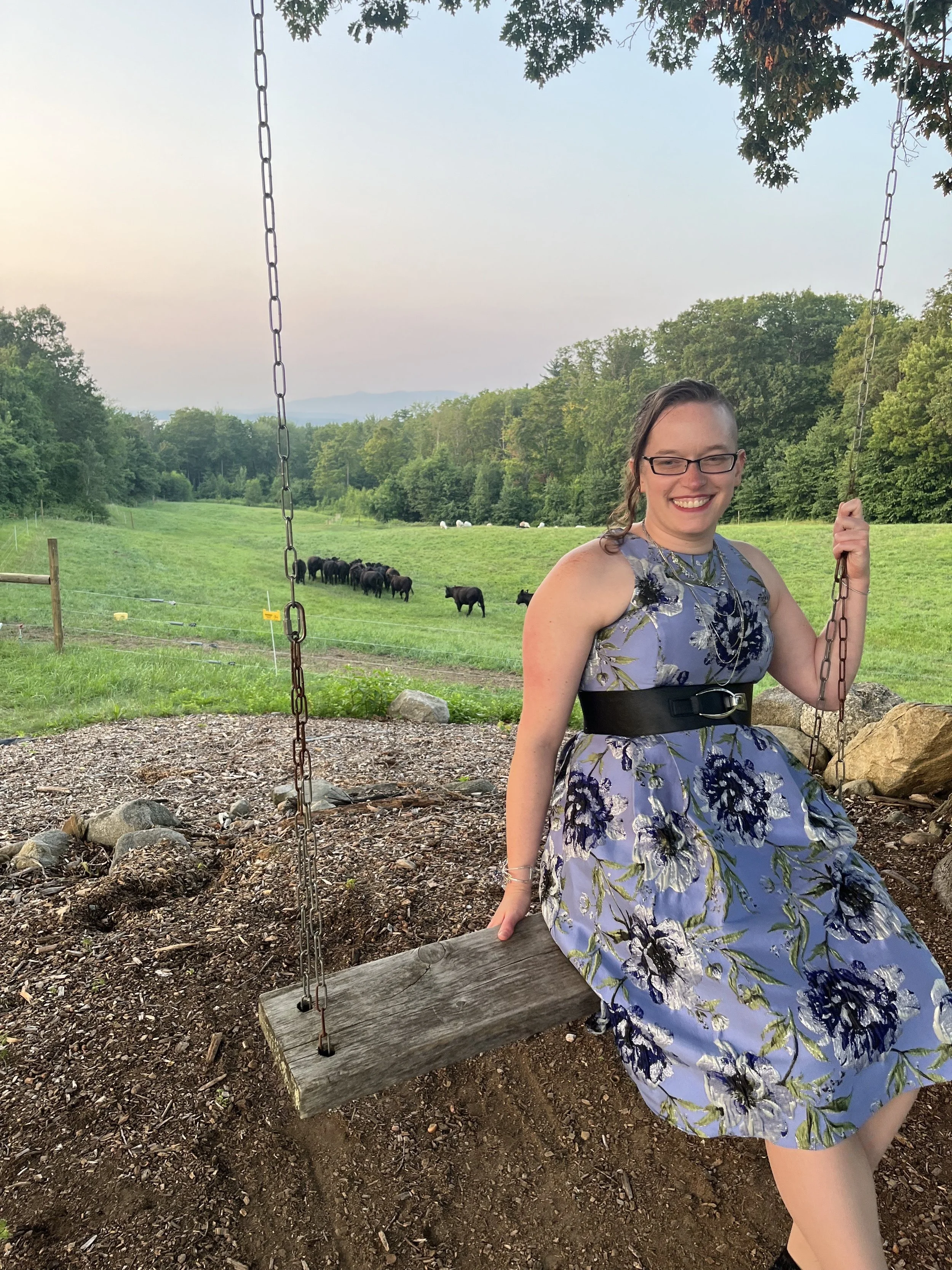 Smiling person sitting on a wooden swing in front of a green pasture at sunset, wearing glasses and a blue floral dress with a black belt. A small herd of cows grazes in the field behind them, with trees and distant hills under a pale sky.