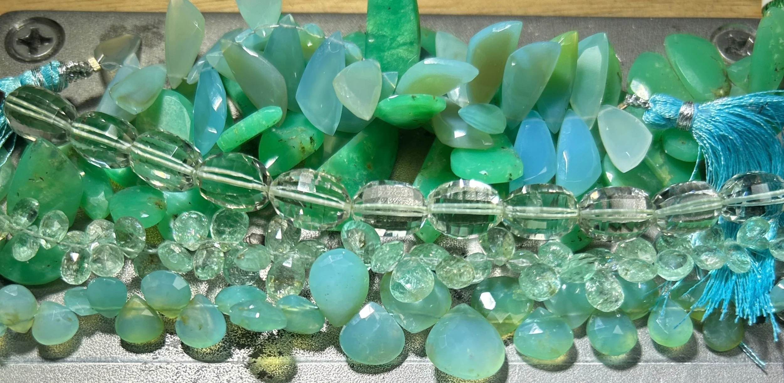 Close-up of green chrysoprase, aqua chalcedony, and clear faceted gemstone strands with turquoise tassels arranged on a workbench.
