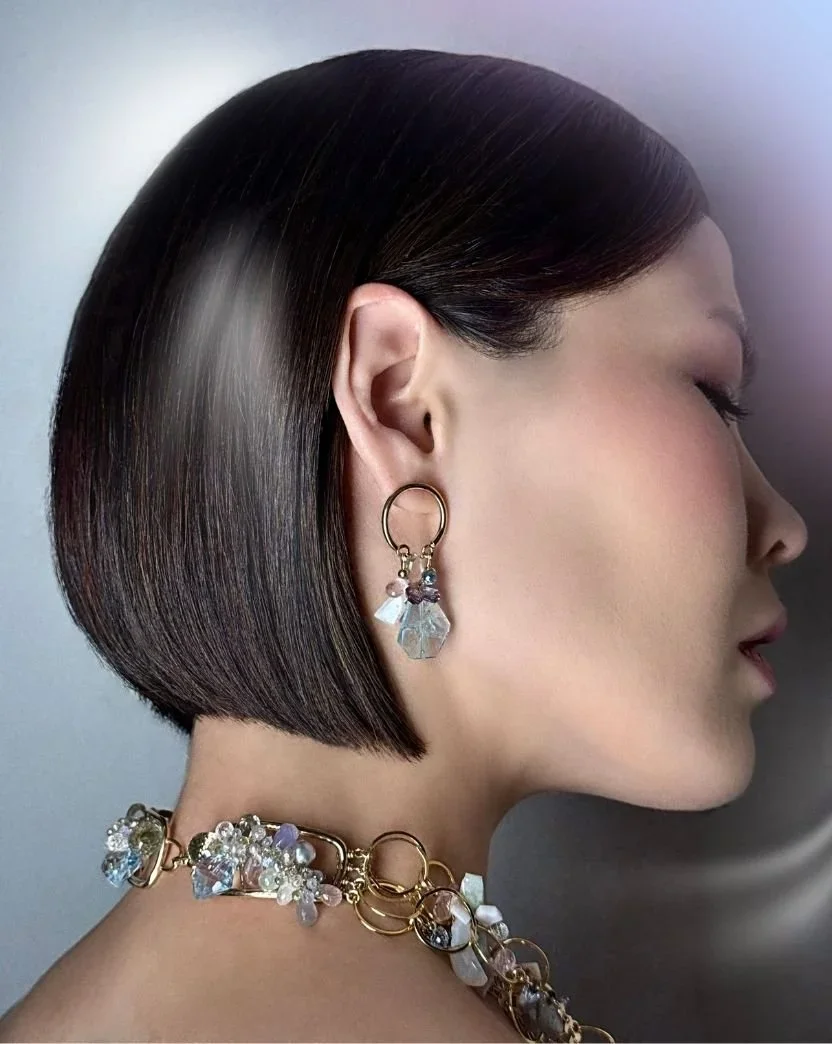 Side-profile portrait of a model with a sleek dark bob wearing a gold hoop earring with pastel gemstone drops and a pastel gemstone collar necklace along the neckline.