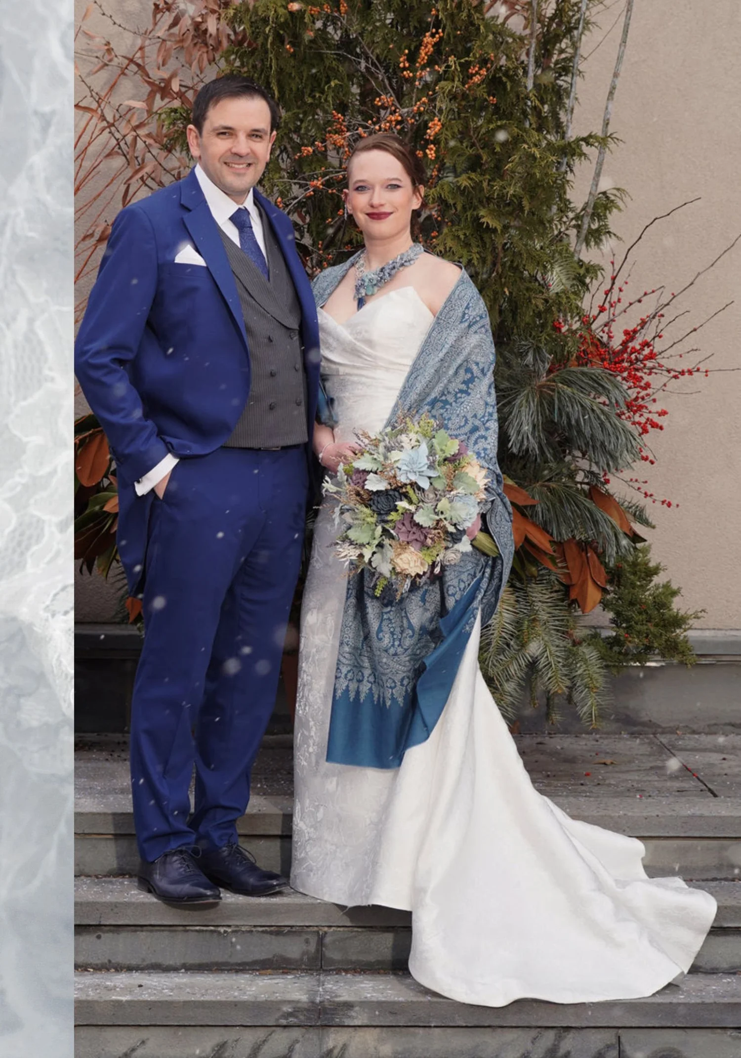 A bride in a strapless ivory gown and a blue paisley shawl, holding a pale bouquet, beside the groom in a blue suit on stone steps, wearing a blue gemstone statement necklace