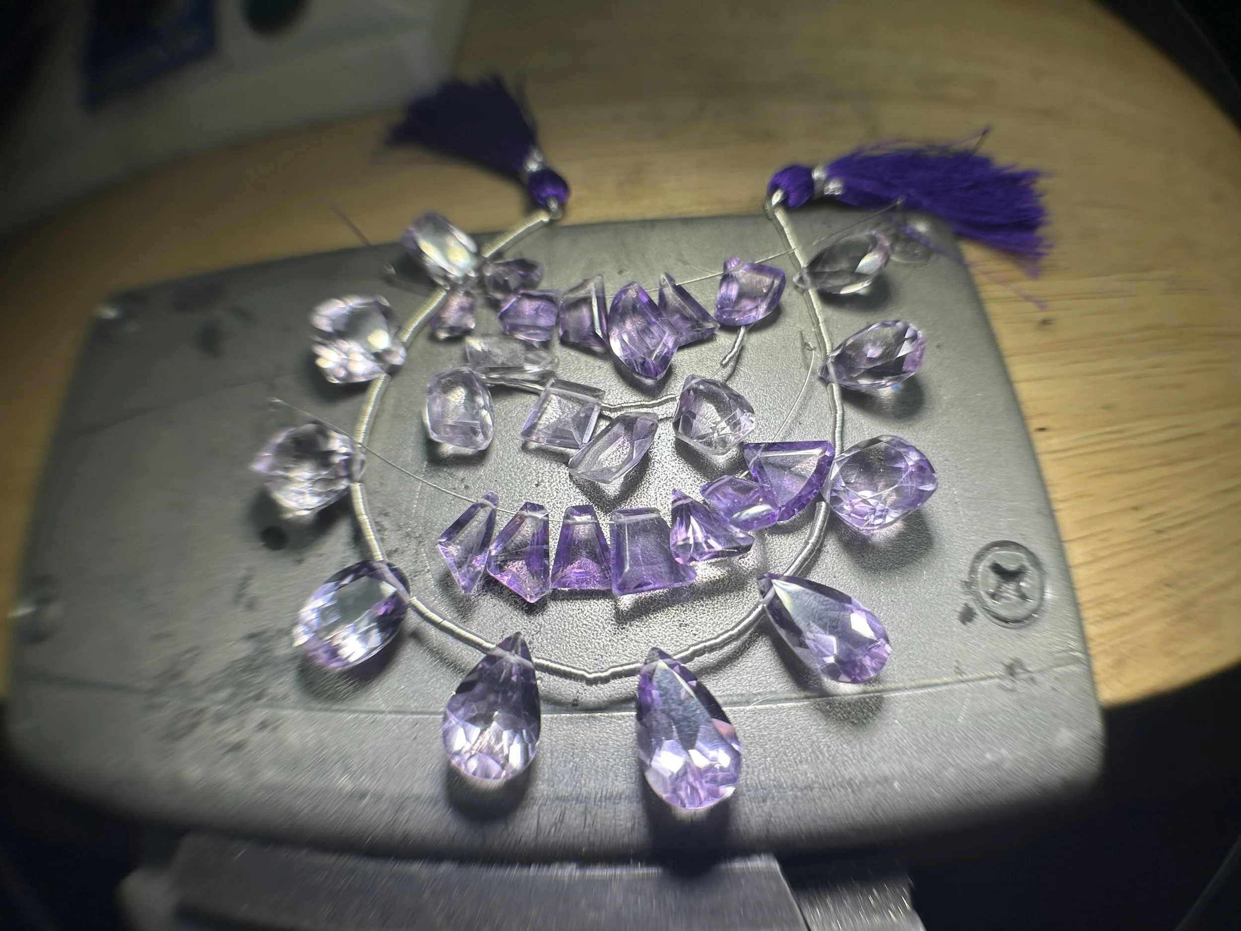 Loose amethyst gemstones in mixed cuts (teardrops, cushions, and trapezoids) arranged on two strands with purple tassels on a metal jeweler’s tray.