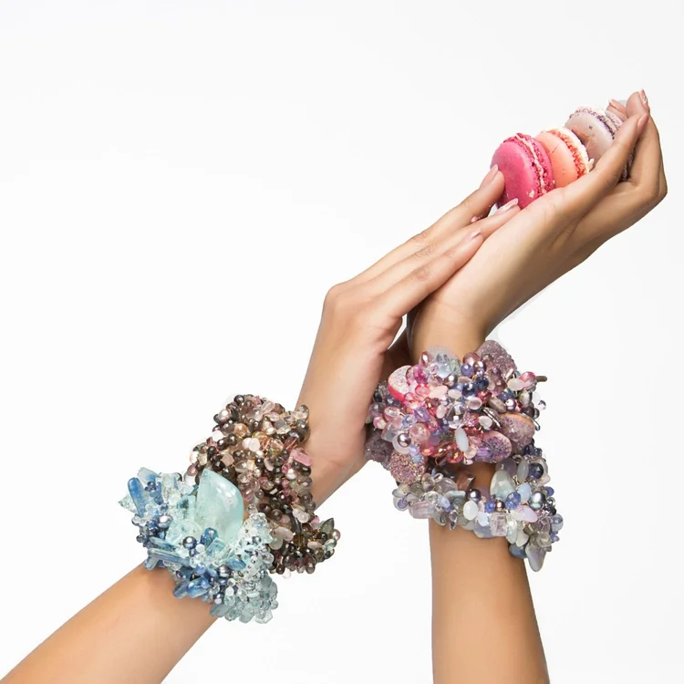 Two raised hands holding macarons, wearing Andrea Li gemstone statement cuffs on both wrists, by Andrea Li