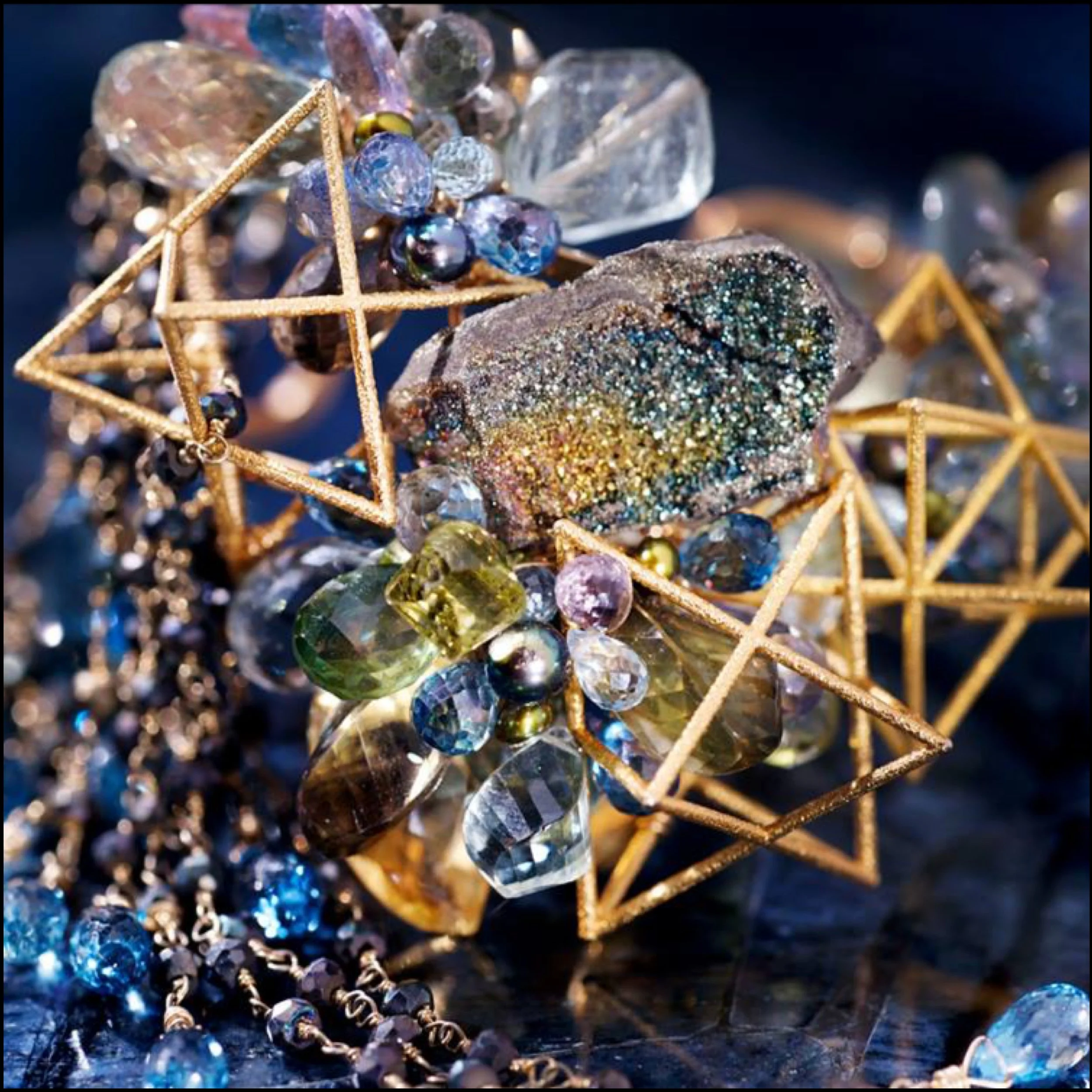 Close-up of a rainbow druzy stone surrounded by colorful faceted gems and gold geometric wire details on a dark surface.