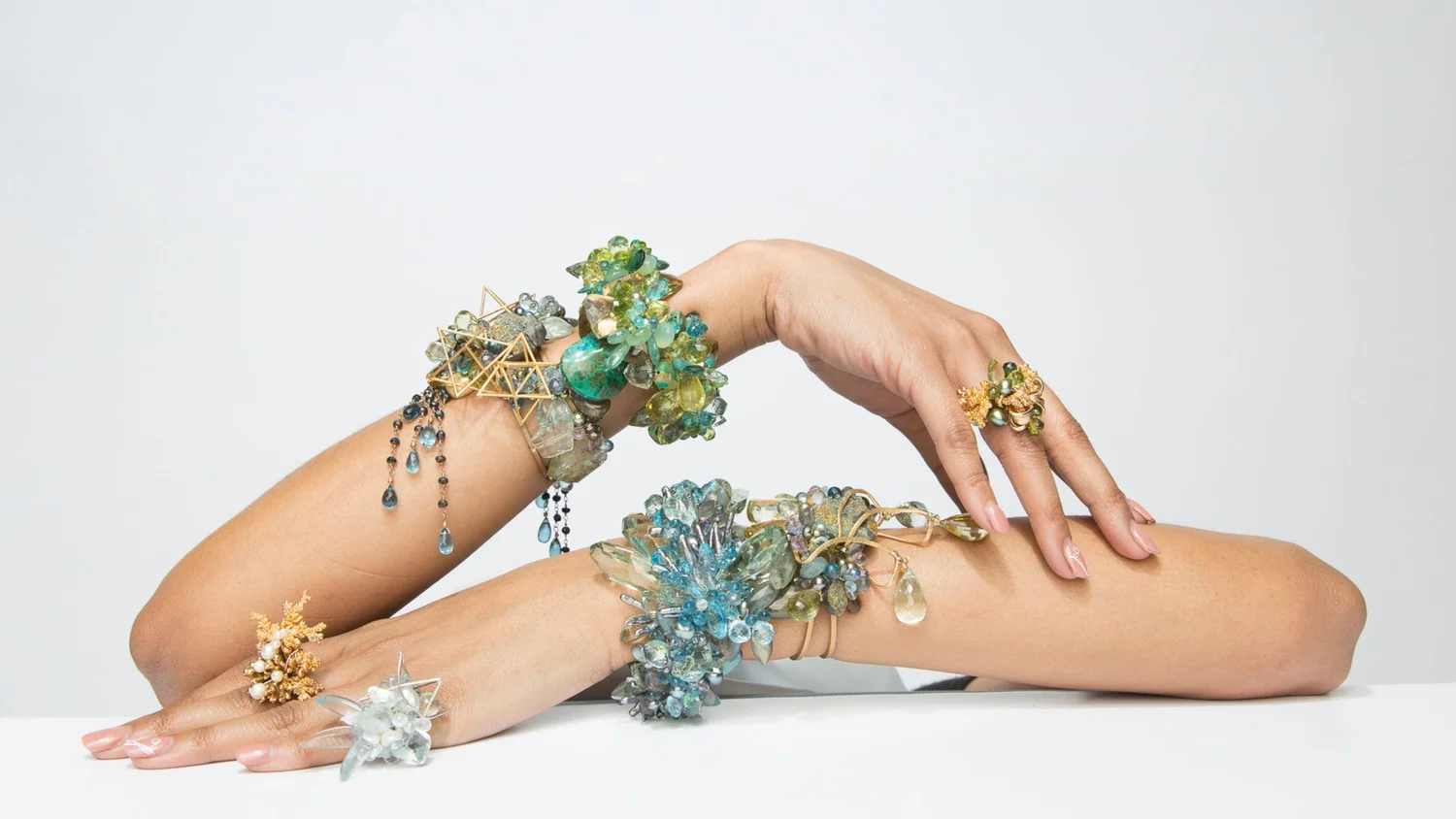 Two arms stacked with Andrea Li statement cuffs and rings: blue and green gemstone clusters, gold wire forms and briolette drops on a white ground