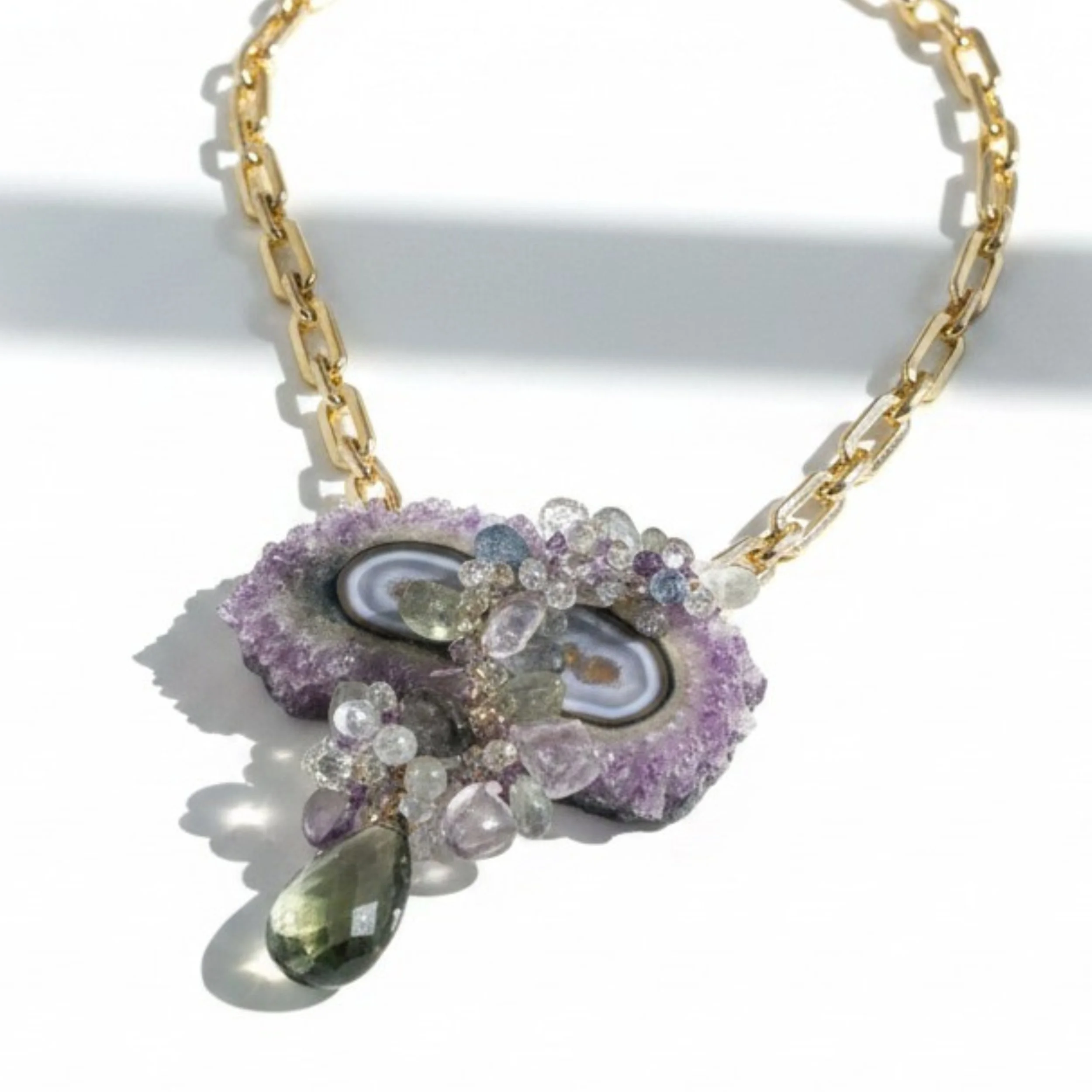 Close-up of a gold paperclip chain necklace with an amethyst stalactite slice pendant and clustered pastel gemstones.