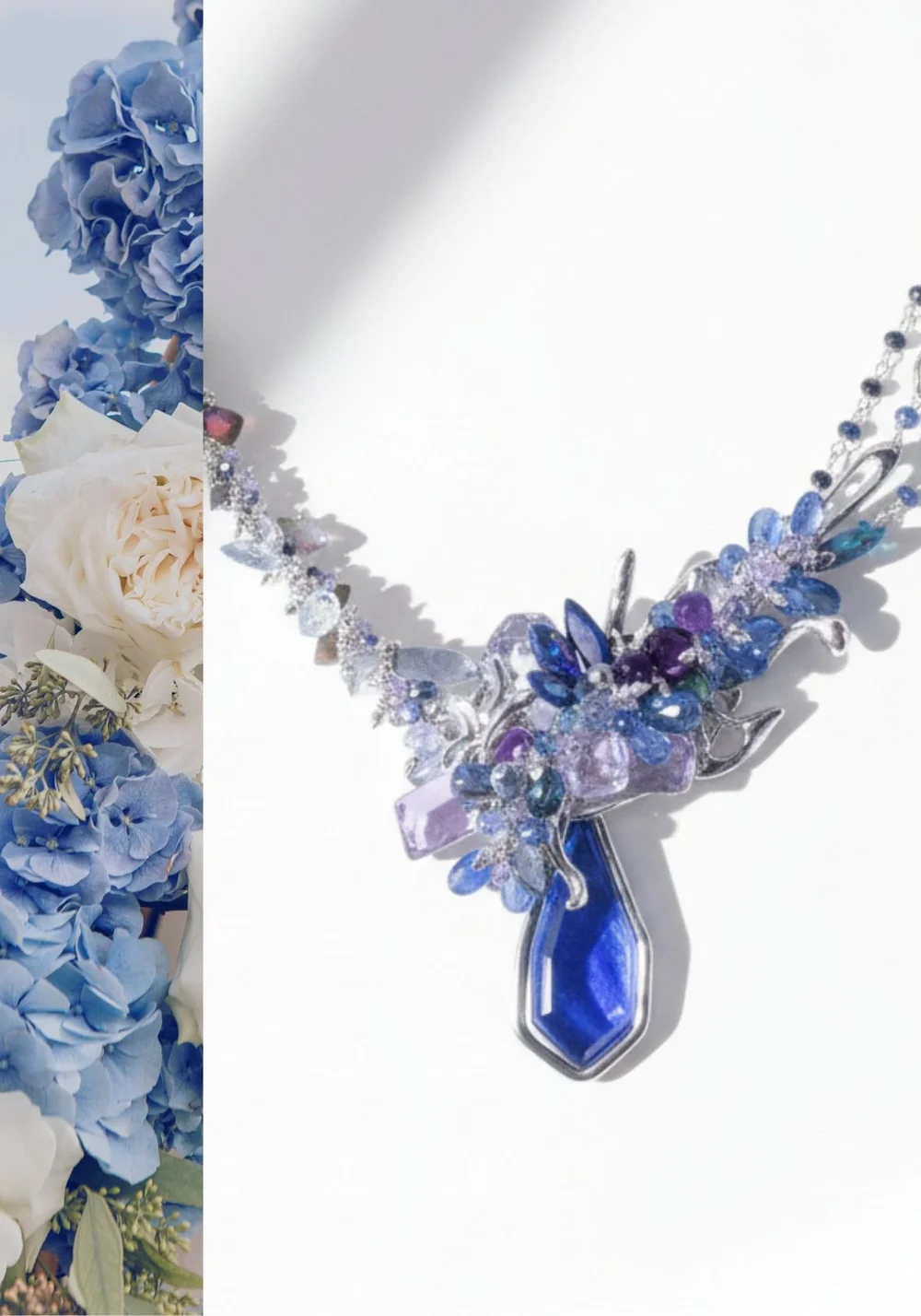 Close-up product photo of a silver statement necklace with layered blue, violet, and clear gemstones forming a floral centerpiece with a bold cobalt pendant, styled beside blue hydrangeas and white roses.