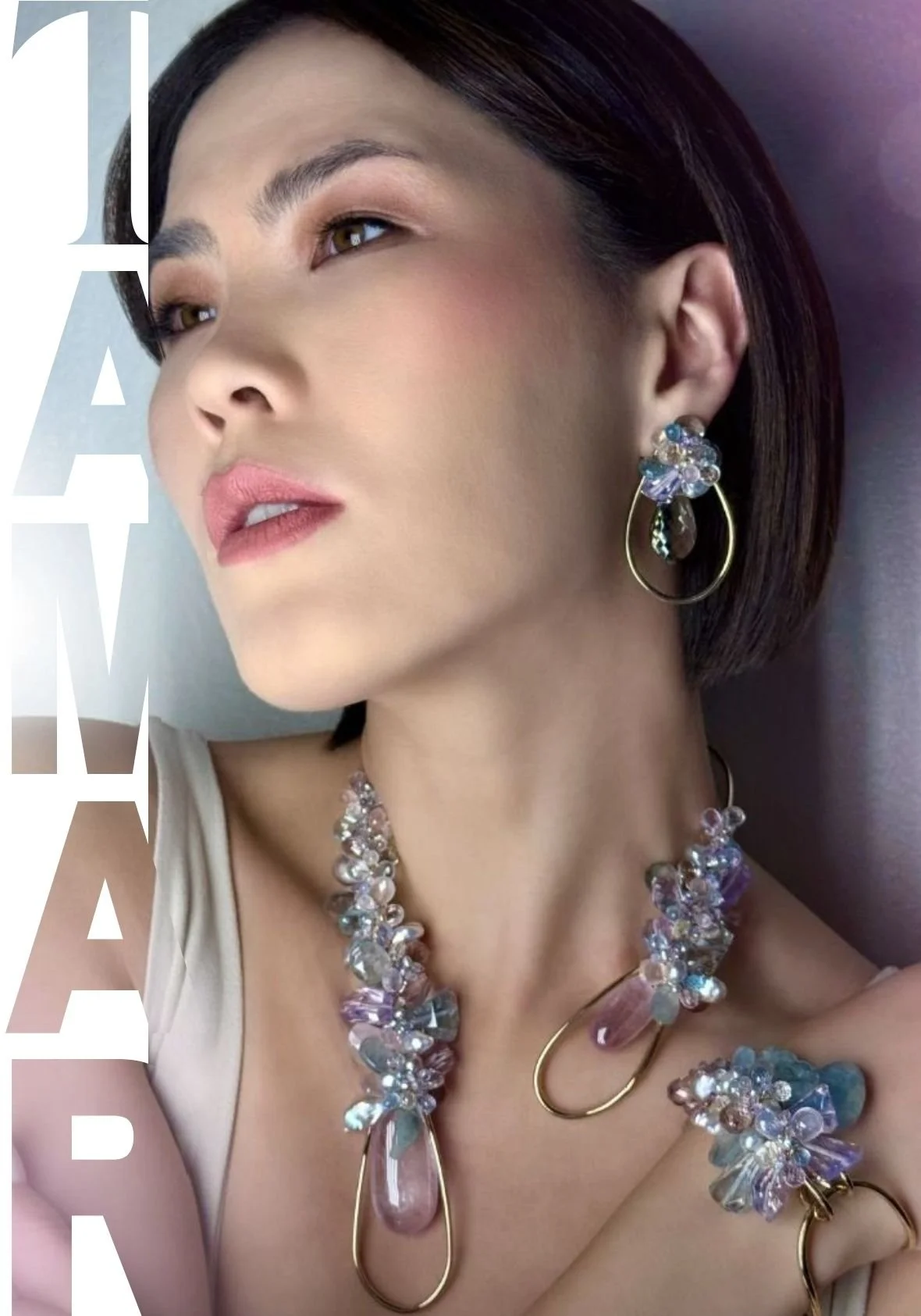 Close-up portrait of a model wearing a pastel gemstone choker necklace with gold hoop elements and a matching gemstone drop earring, with the word "TAMAR" printed vertically along the left edge.