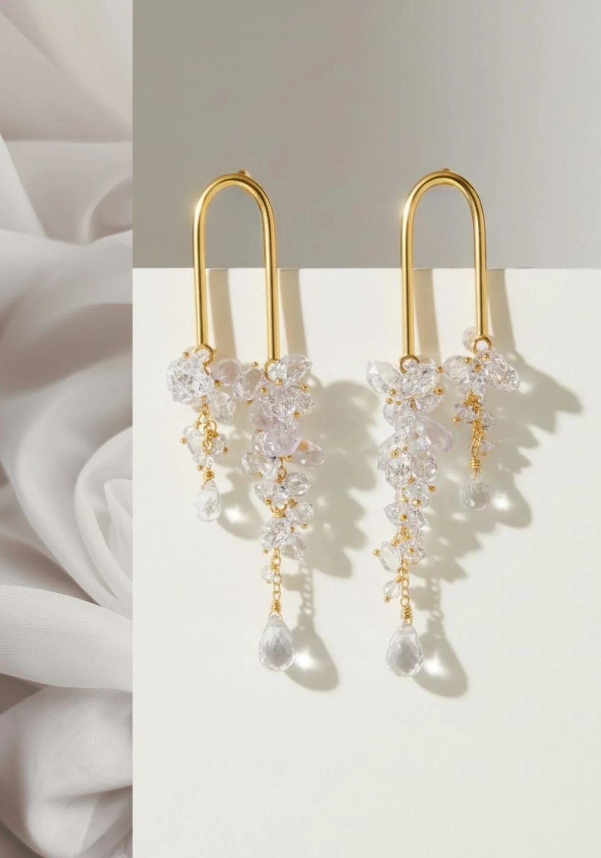 Pair of gold bridal earrings with clustered clear crystals and dangling teardrop accents, displayed on a white pedestal beside softly draped fabric.