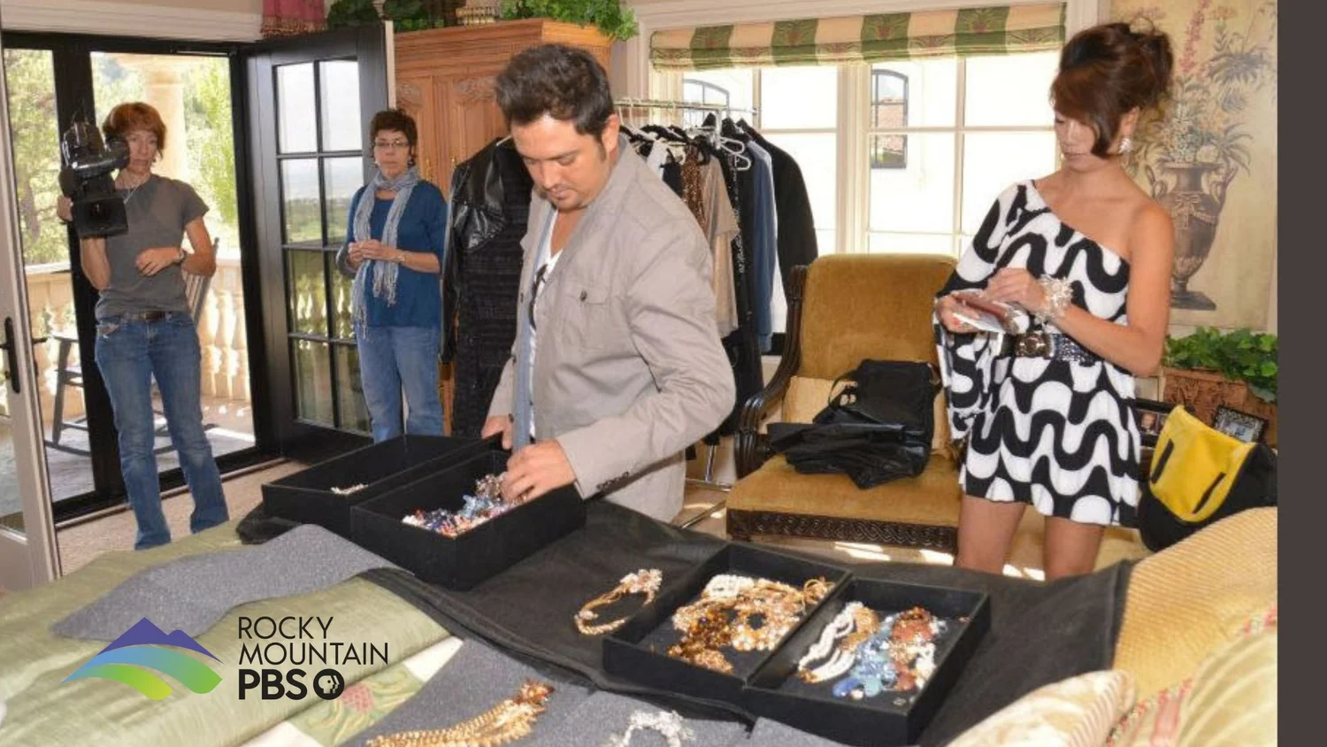 Behind the scenes of the Rocky Mountain PBS Arts District shoot: fashion designer Gino Velardi sorts trays of Andrea Li jewelry on a bed while Andrea, in a black and white dress, and the camera crew look on