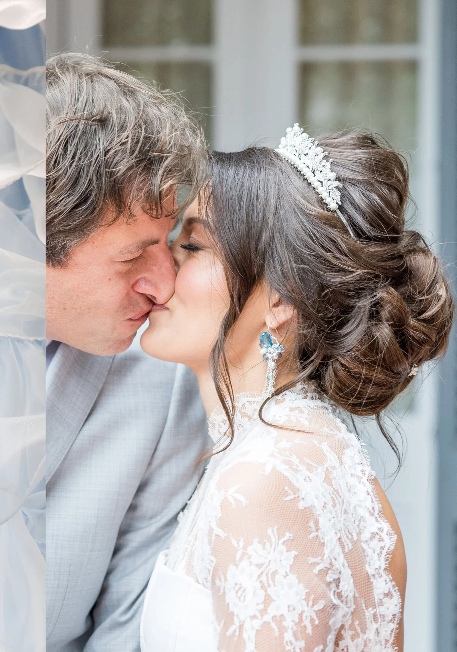 A groom in a gray suit kissing a bride in a jeweled tiara and a high-neck lace gown, blue gemstone drop earrings at her ear