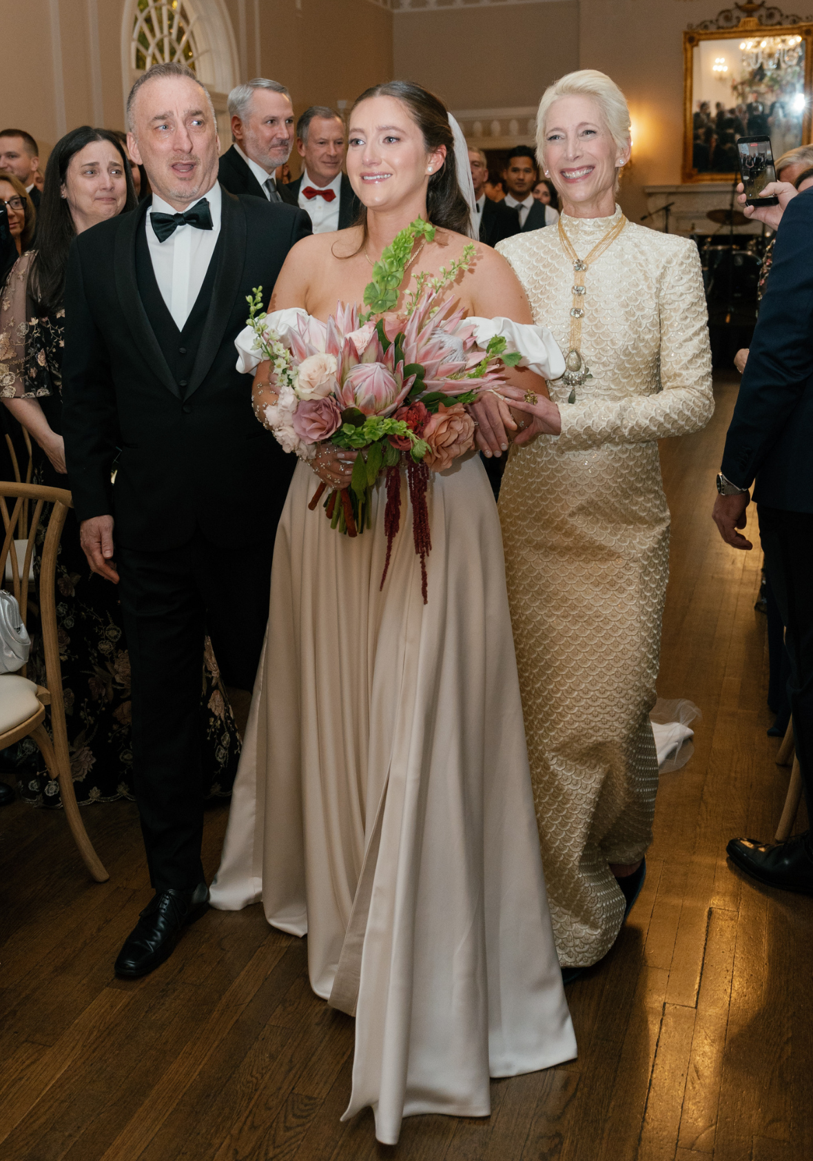 Bride walking down the aisle indoors holding a large pink and blush bouquet, flanked by a man in a black tuxedo and a smiling woman in a long cream textured gown wearing a statement necklace.